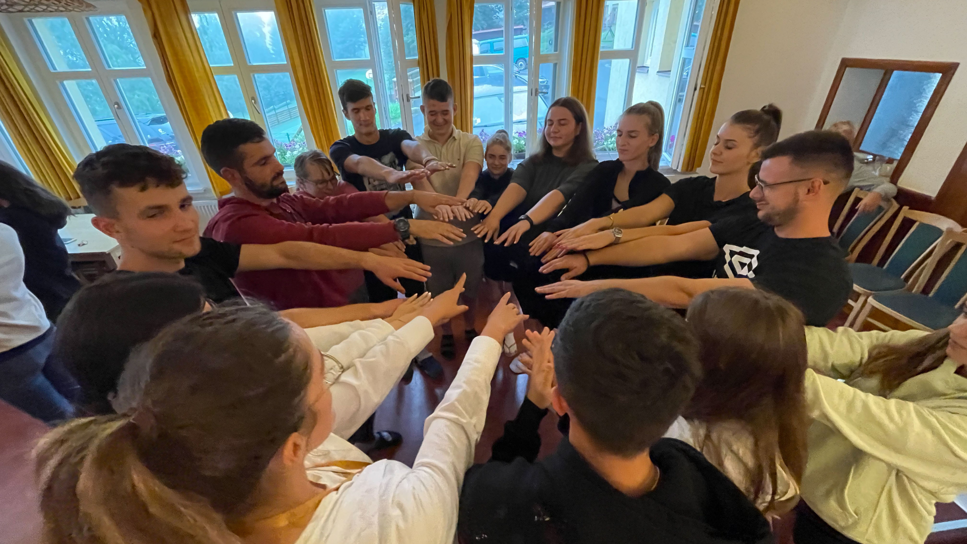 Group of people standing in a circle with outstretched arms, participating in a team-building activity indoors near large windows.