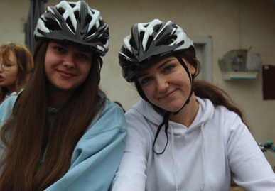 Two young women wearing bike helmets and casual clothing, smiling at the camera in an indoor setting.