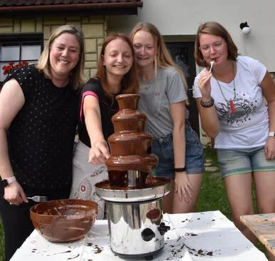 Four young women smiling and posing around a chocolate fountain outdoors.