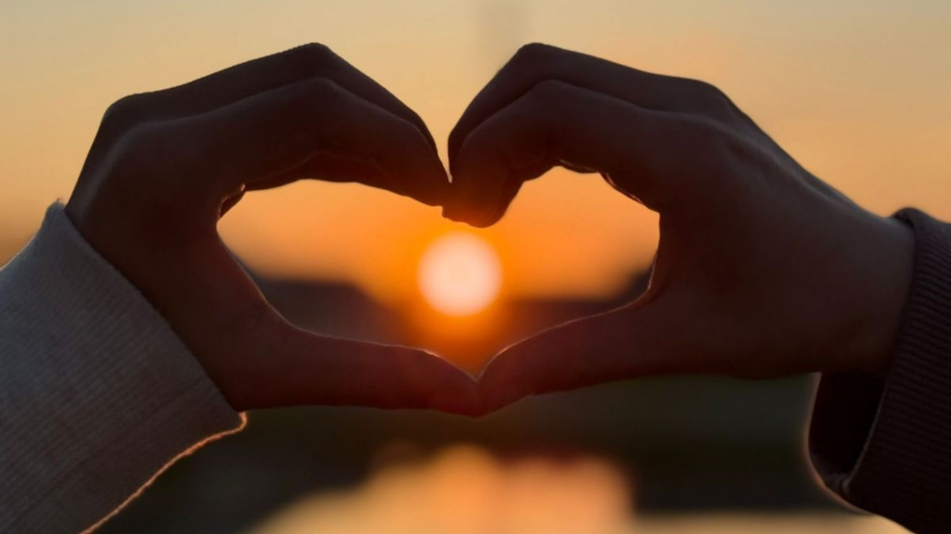 Hands forming a heart shape around a setting sun at sunset.