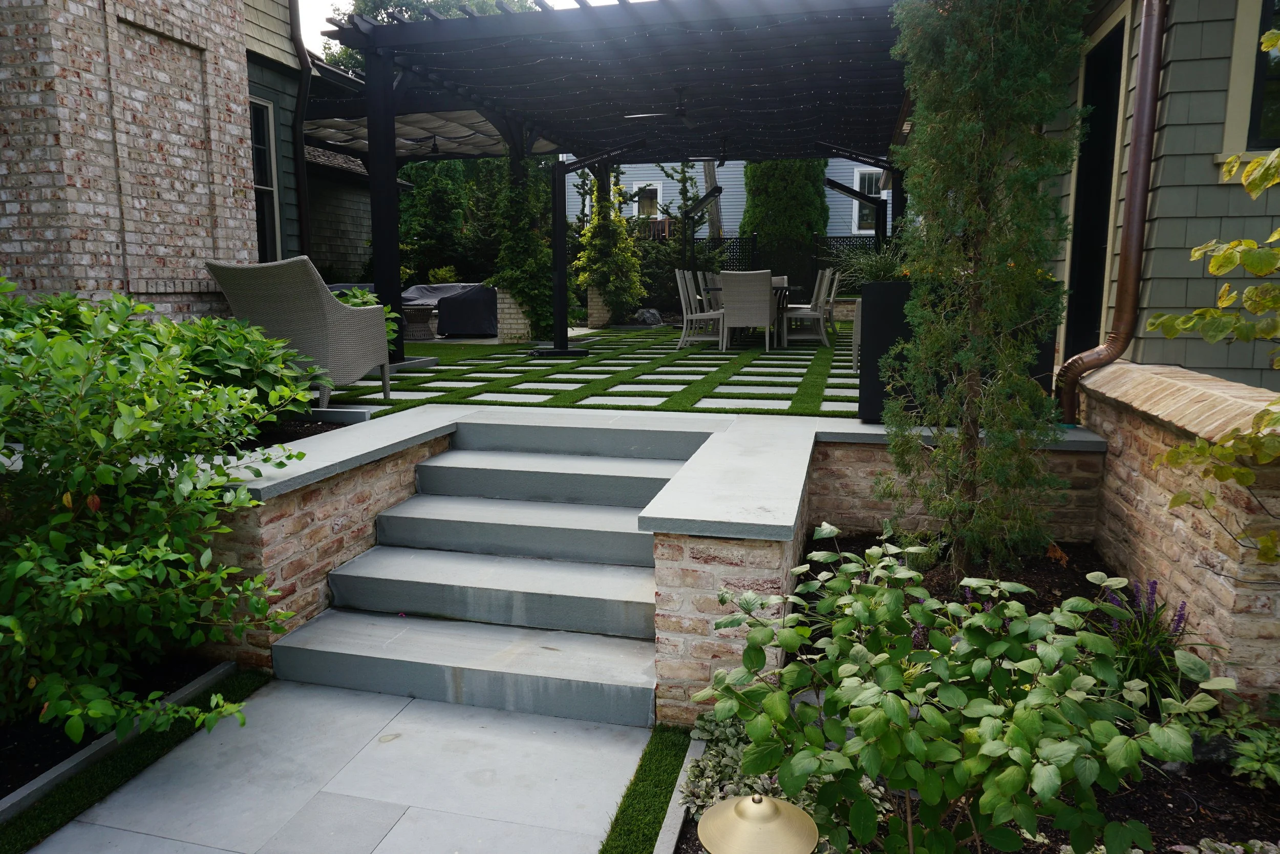 A backyard patio with a concrete staircase leading up to a garden area with a grass and stone checkered pattern. There are outdoor chairs and a table under a dark pergola, surrounded by green plants and shrubbery. A section of a house with brick and siding is visible.