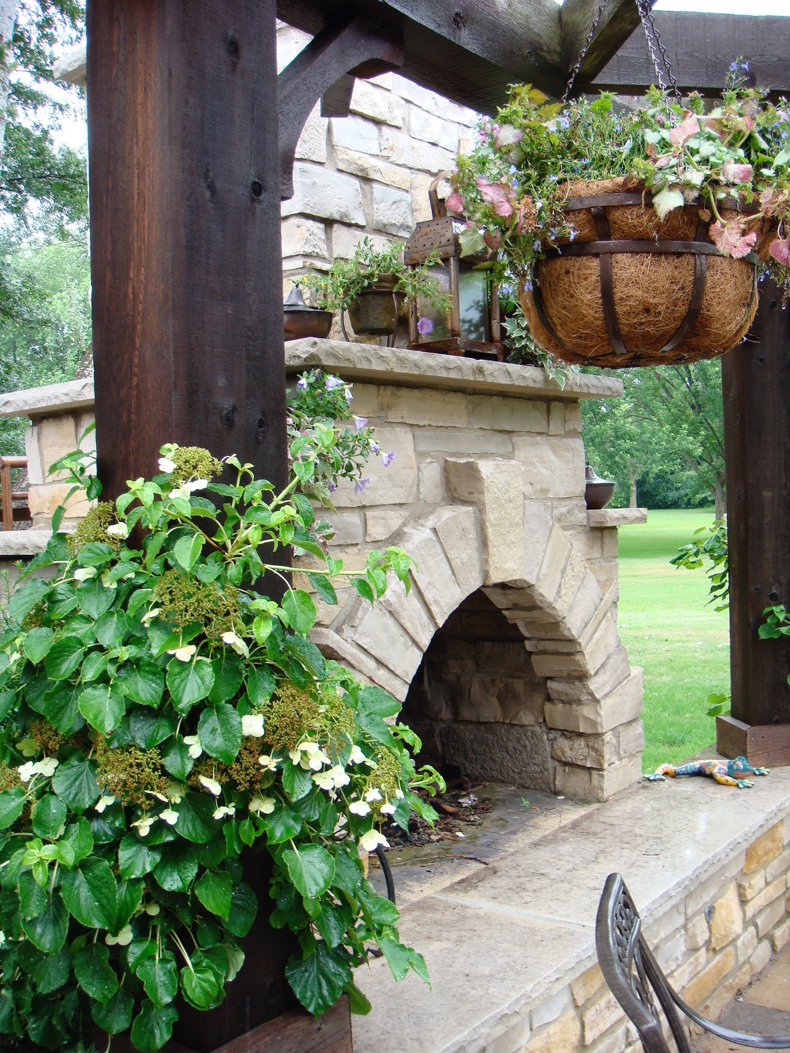 A stone outdoor fireplace with a wooden mantel, surrounded by lush green plants and a hanging flower basket.