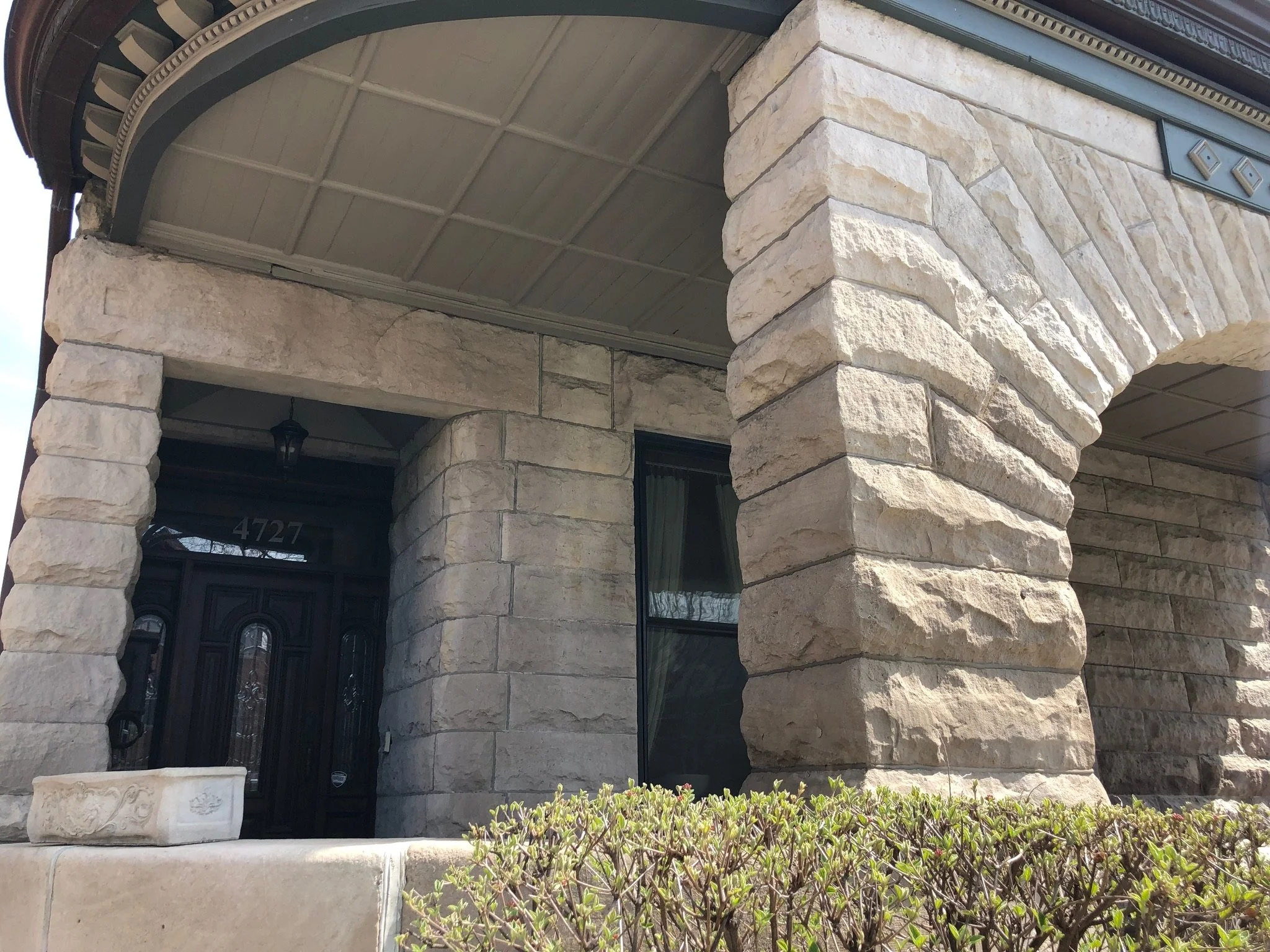 Close-up of a house's front porch with stone columns, a decorative stone flower pot, and a black front door. The porch ceiling is painted in a neutral tone with visible grid pattern.