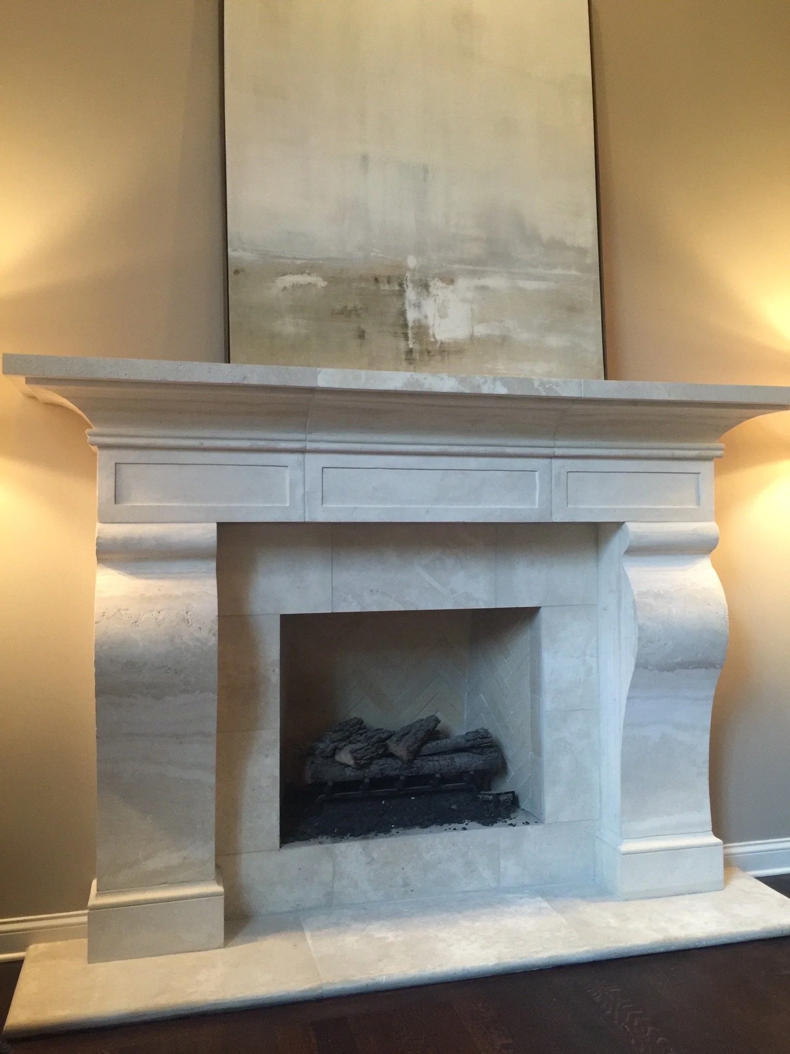 A white marble fireplace with a mantel and an abstract painting hanging above it, with a wood and stone floor.