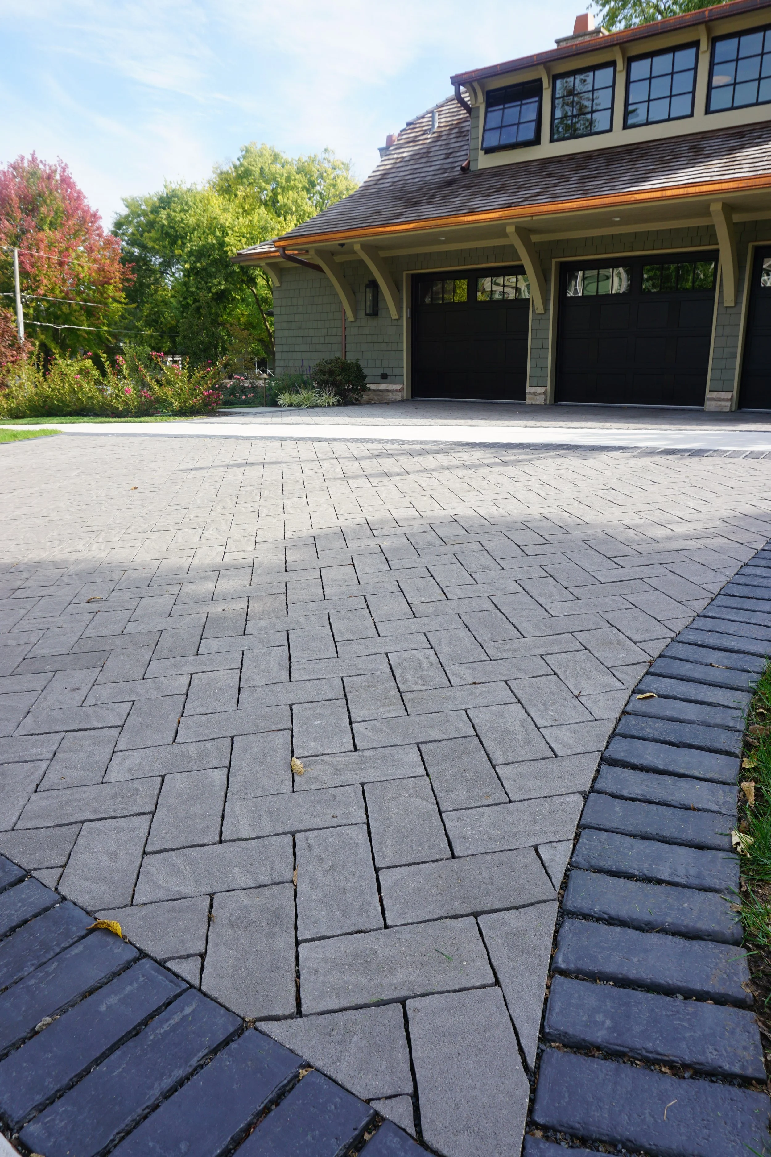 A residential house with a two-car garage, gray brick exterior, brown shingle roof, and landscaping with bushes and colorful trees. Paved driveway and walkway in front.