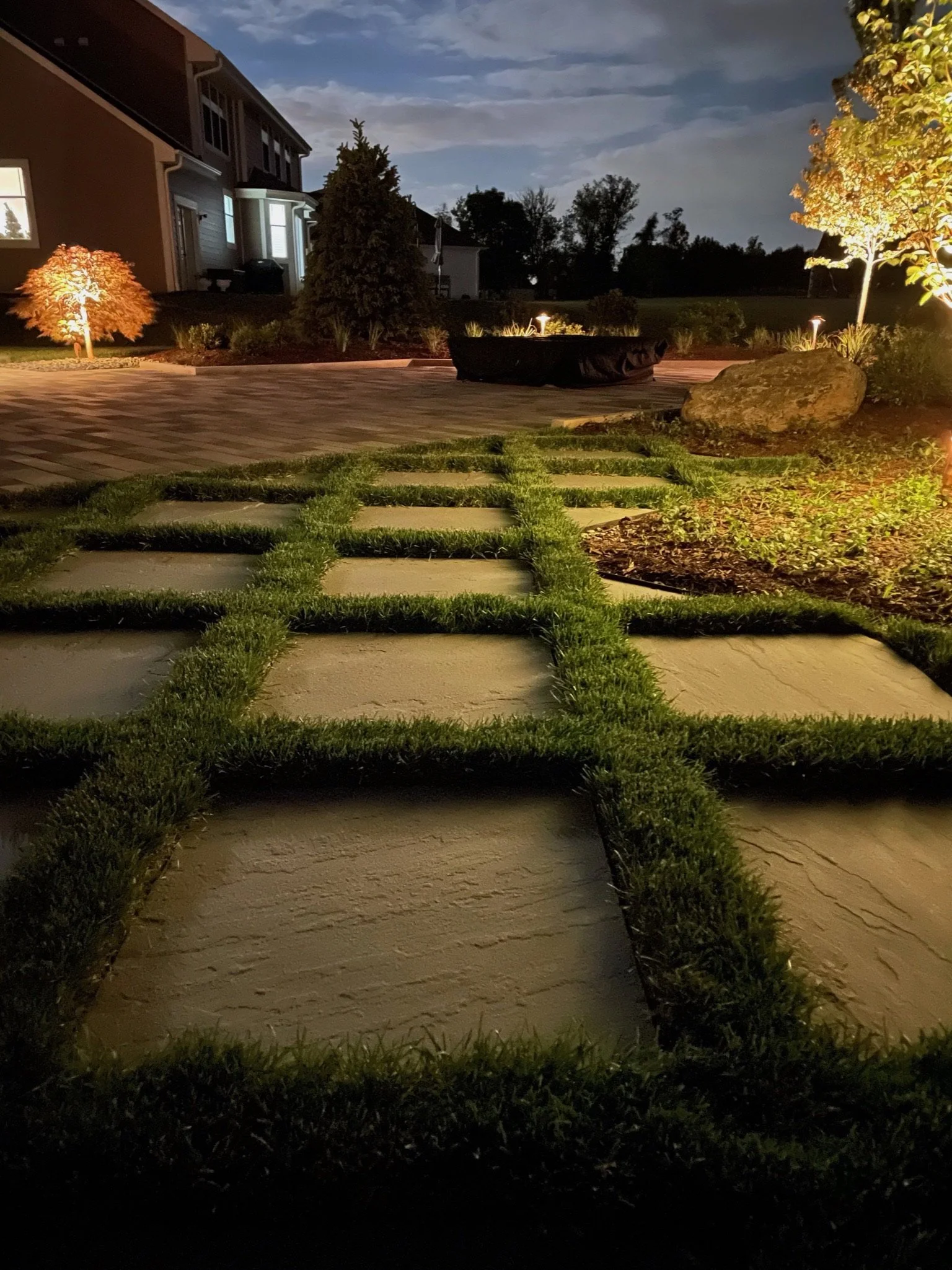 Nighttime backyard with illuminated trees, shrubs, and pathway leading to a house with windows, under a cloudy sky.