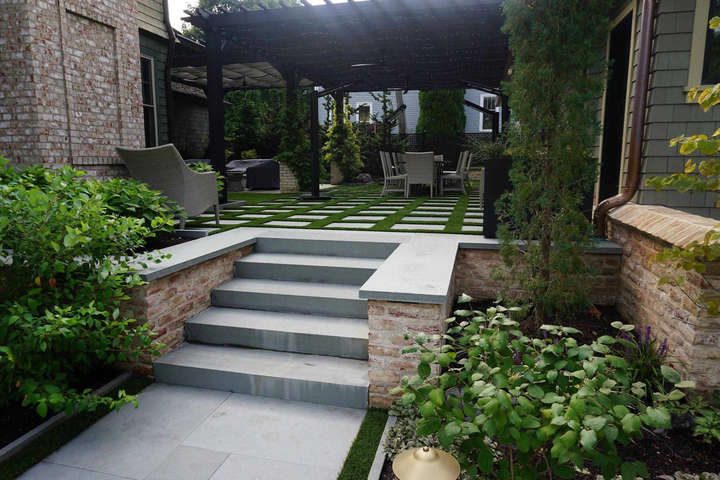 A backyard patio with concrete steps leading up to a raised area with outdoor dining and garden space, surrounded by greenery and brick walls.