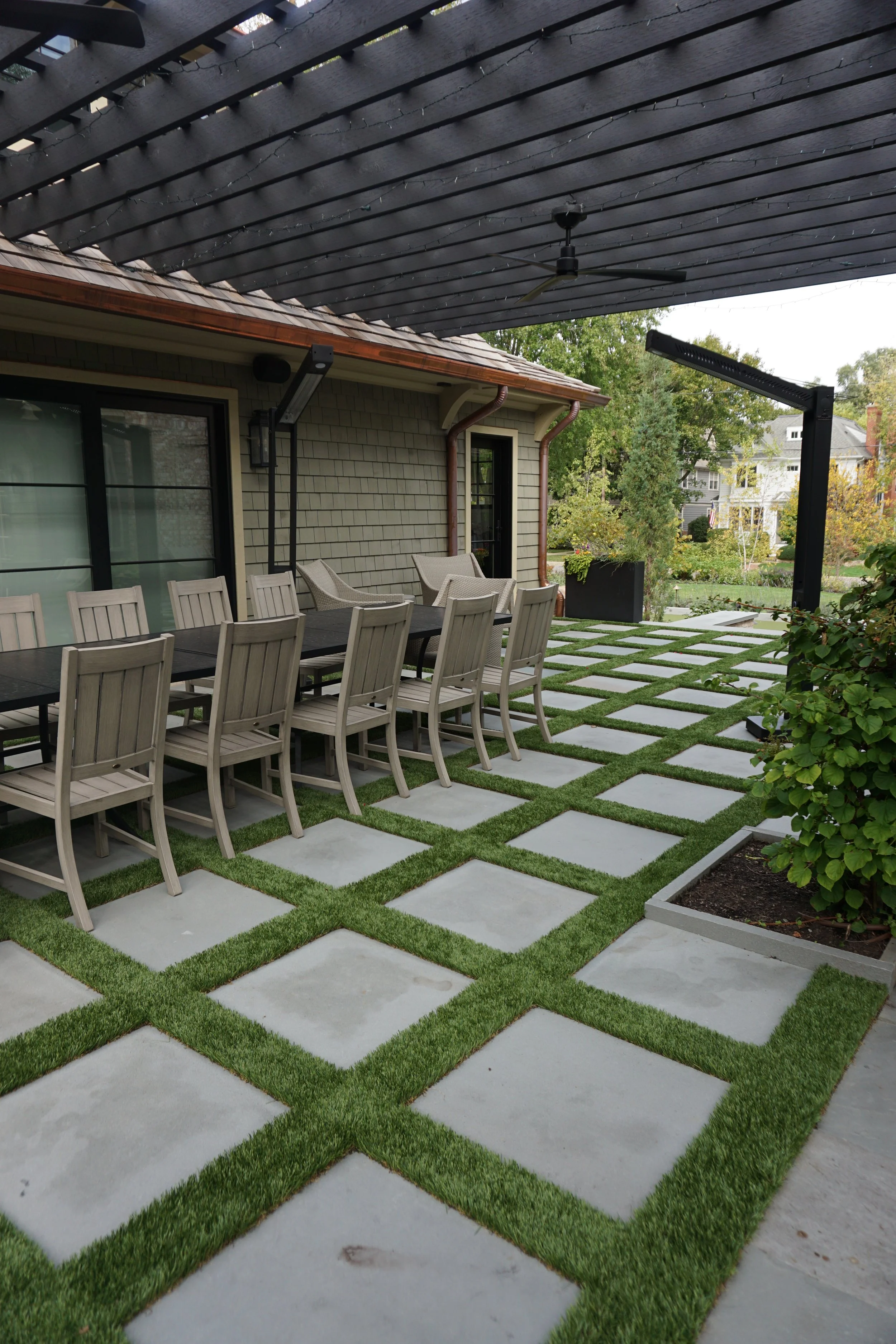 An outdoor patio area with a long dining table and 10 chairs under a black wooden pergola with string lights, adjacent to a house with gray shingle siding; the patio has a pattern of concrete pavers with grass in between, surrounded by trees and neighboring houses.