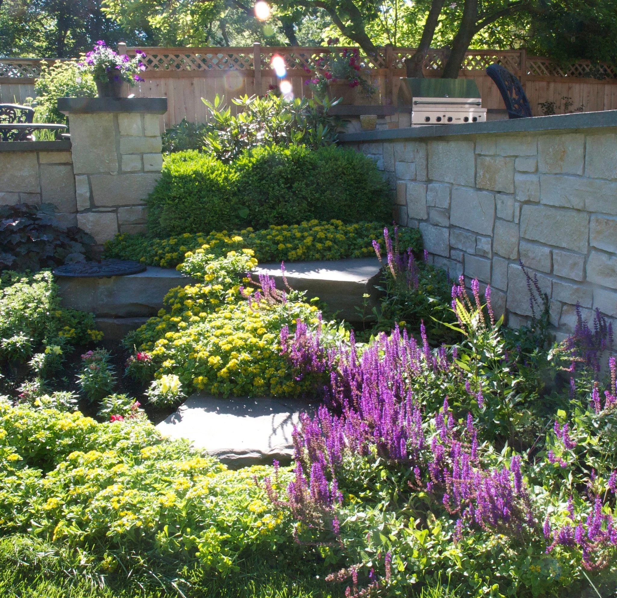 A backyard garden with various colorful flowers, green plants, and shrubs in front of a stone wall. There is a wooden fence with lattice on top, a barbecue grill, and outdoor seating visible in the background, surrounded by trees and sunlight.