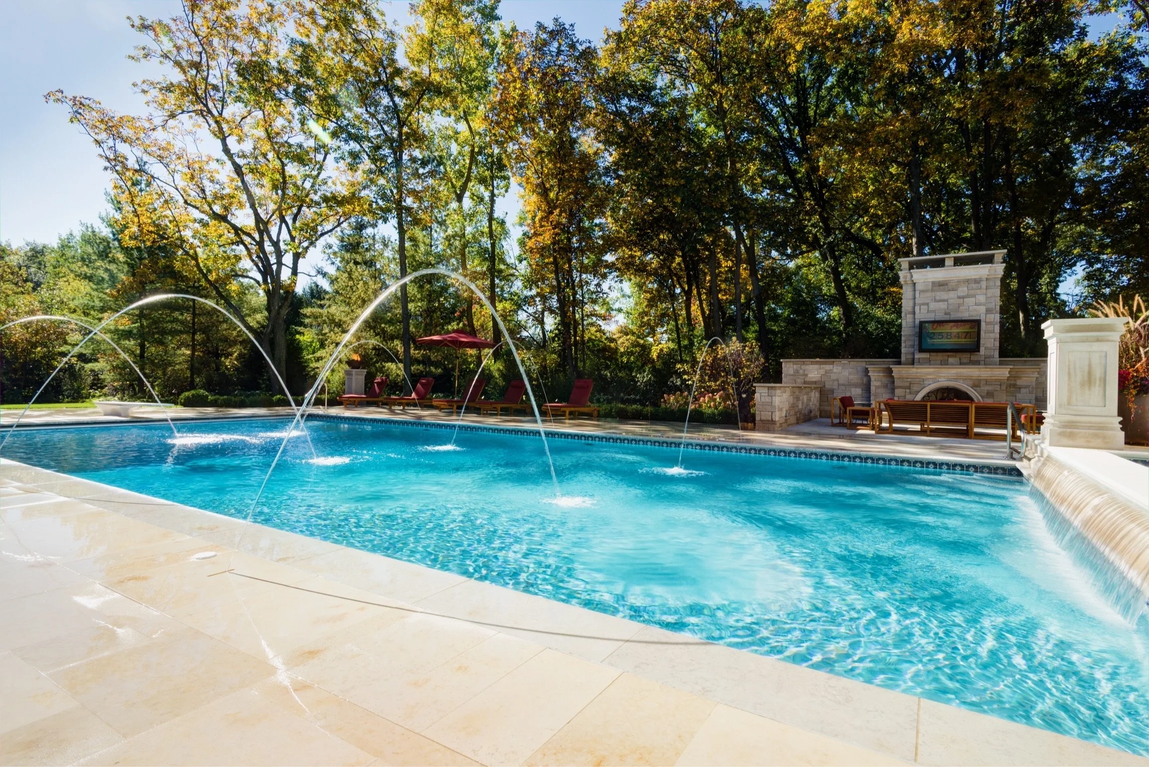 Inground swimming pool with water jets and fountain features, surrounded by poolside chairs, a stone fireplace with a TV, and trees with fall foliage under a clear blue sky.