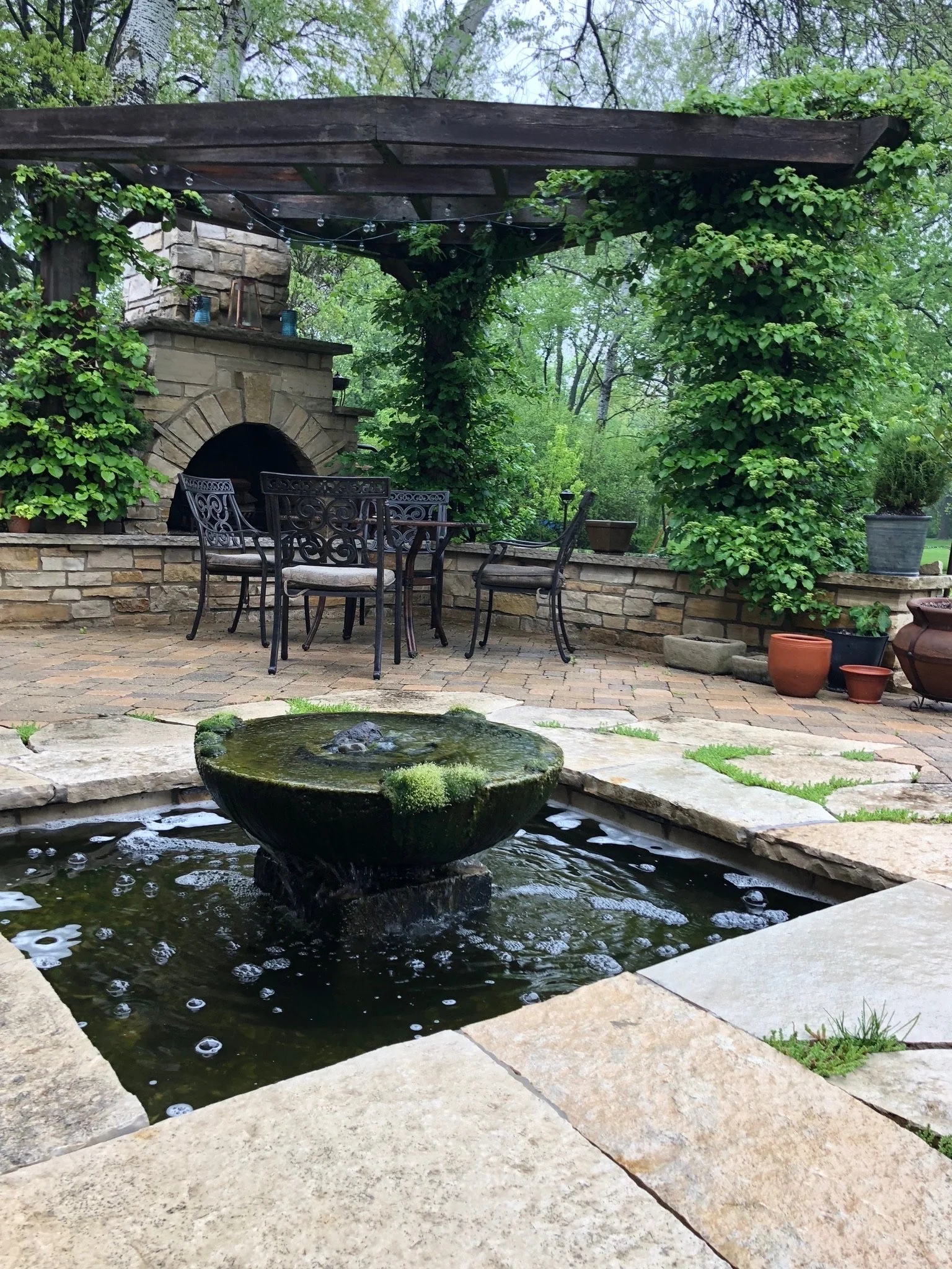 A backyard with a stone patio, a water fountain, surrounded by lush green trees and plants, with outdoor furniture and string lights hanging from a wooden pergola.