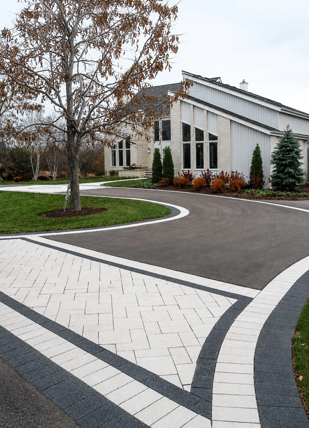 A modern house with large windows and a sloped roof, surrounded by a landscaped yard with trees, shrubs, and a curved driveway with white, gray, and black pavers.