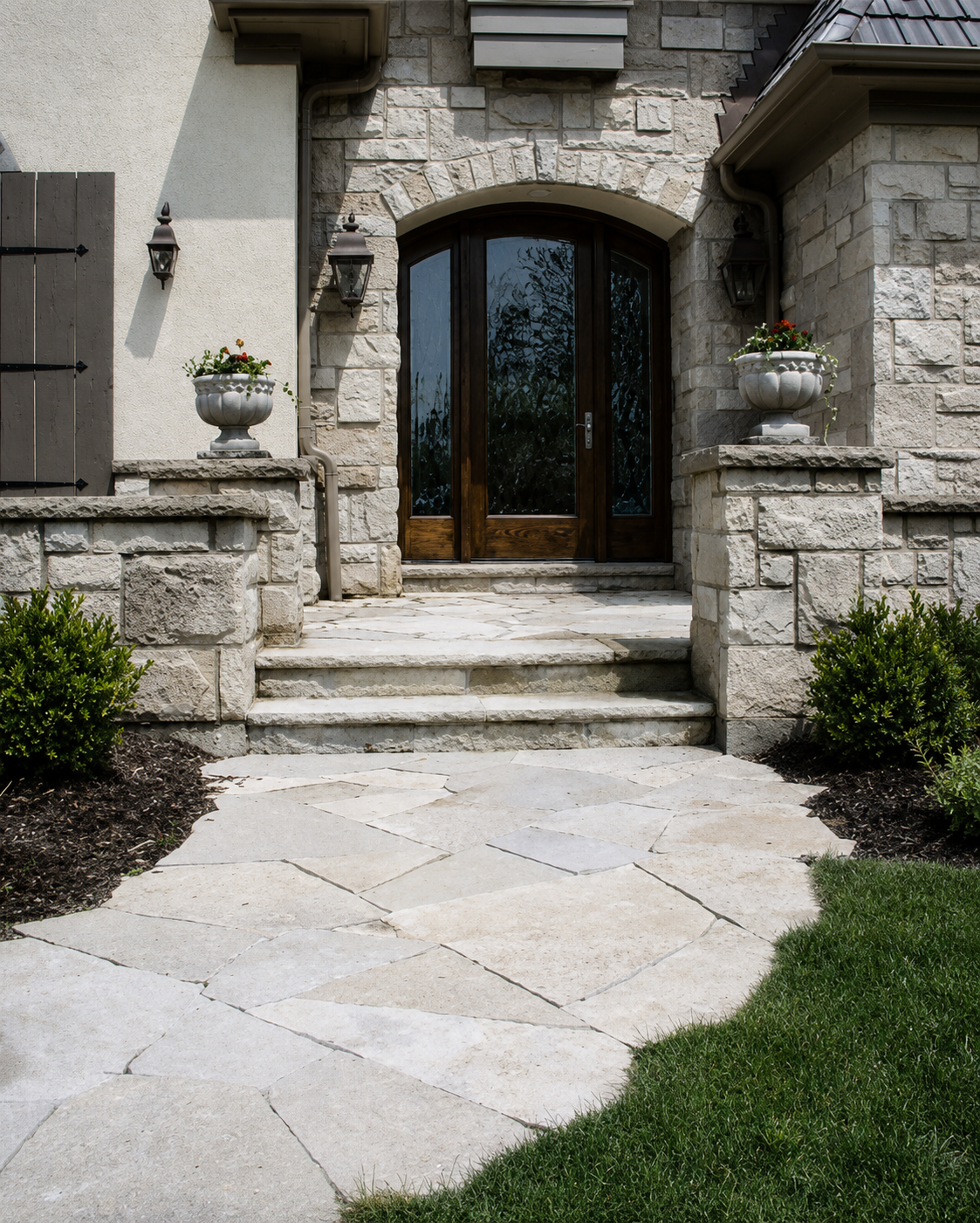 Front entrance of a house with a wooden door, stone steps, two white decorative planters with red and white flowers, wall-mounted lanterns, and a stone walkway leading to the steps.