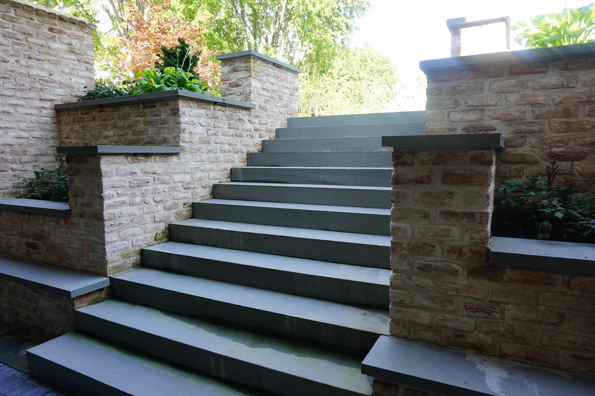 Exterior staircase with modern gray steps, brick walls on sides, and lush green trees in the background.