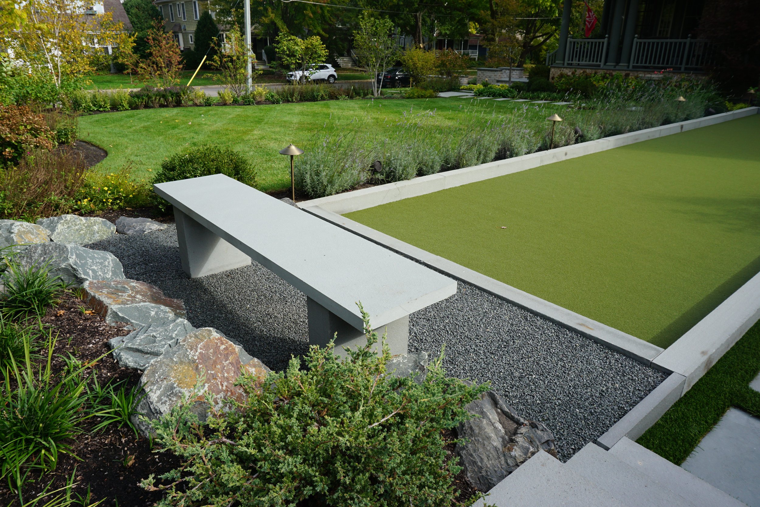 A backyard garden with a small lawn area, flower bushes, and a practice putting green. There are landscape lights, rocks, and gravel edging, with houses and trees in the background.