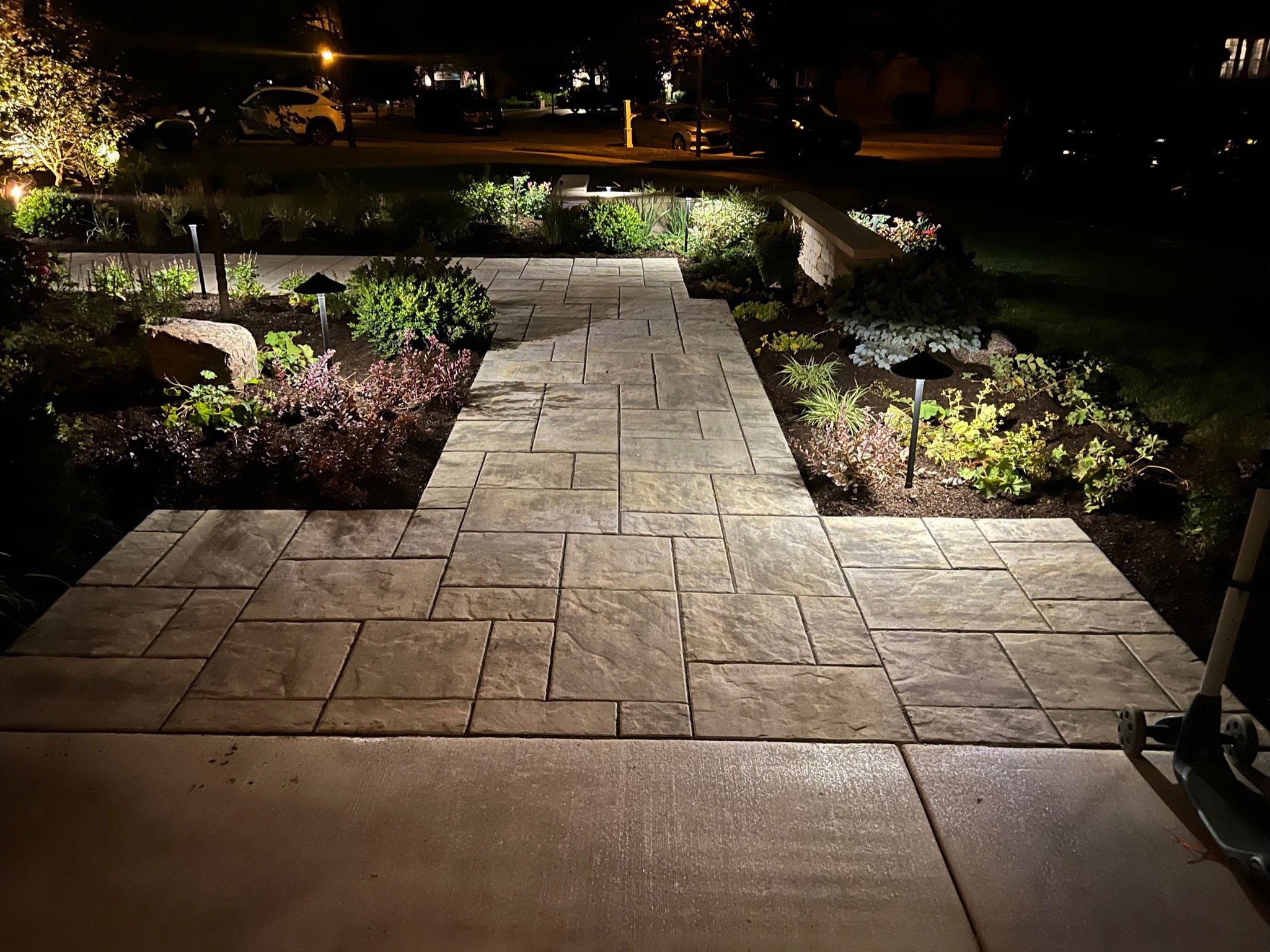 Nighttime view of a backyard patio with a stone pathway, garden beds with plants and small lights along the edges, and a lawn beyond, with parked cars and houses visible in the background across the street.