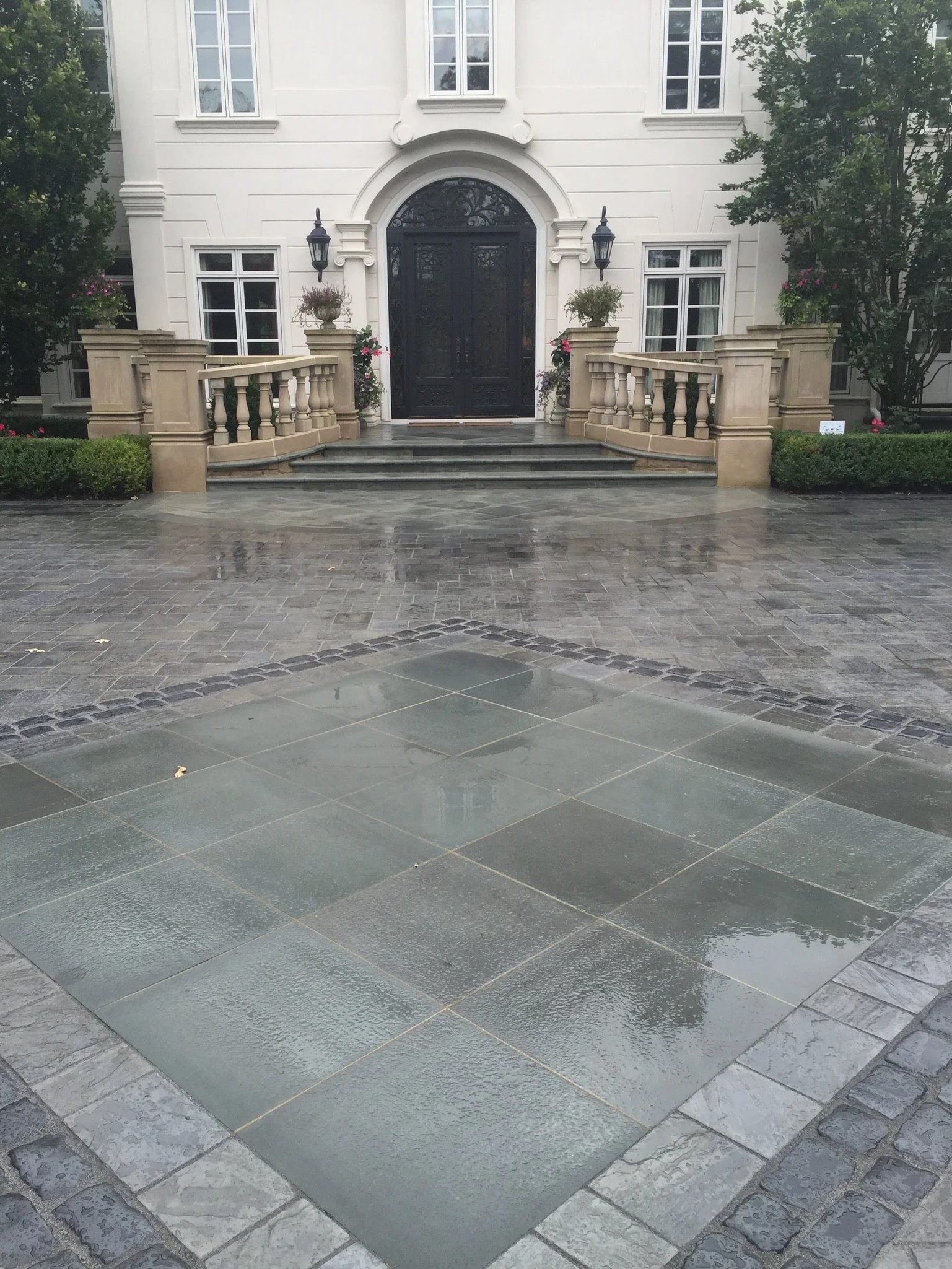 The image shows a front yard with a patio paved with large tiles, leading up to a white house with a decorative black wrought iron front door, flanked by two outdoor lanterns, and decorated with potted plants and flowers on either side of the entranc