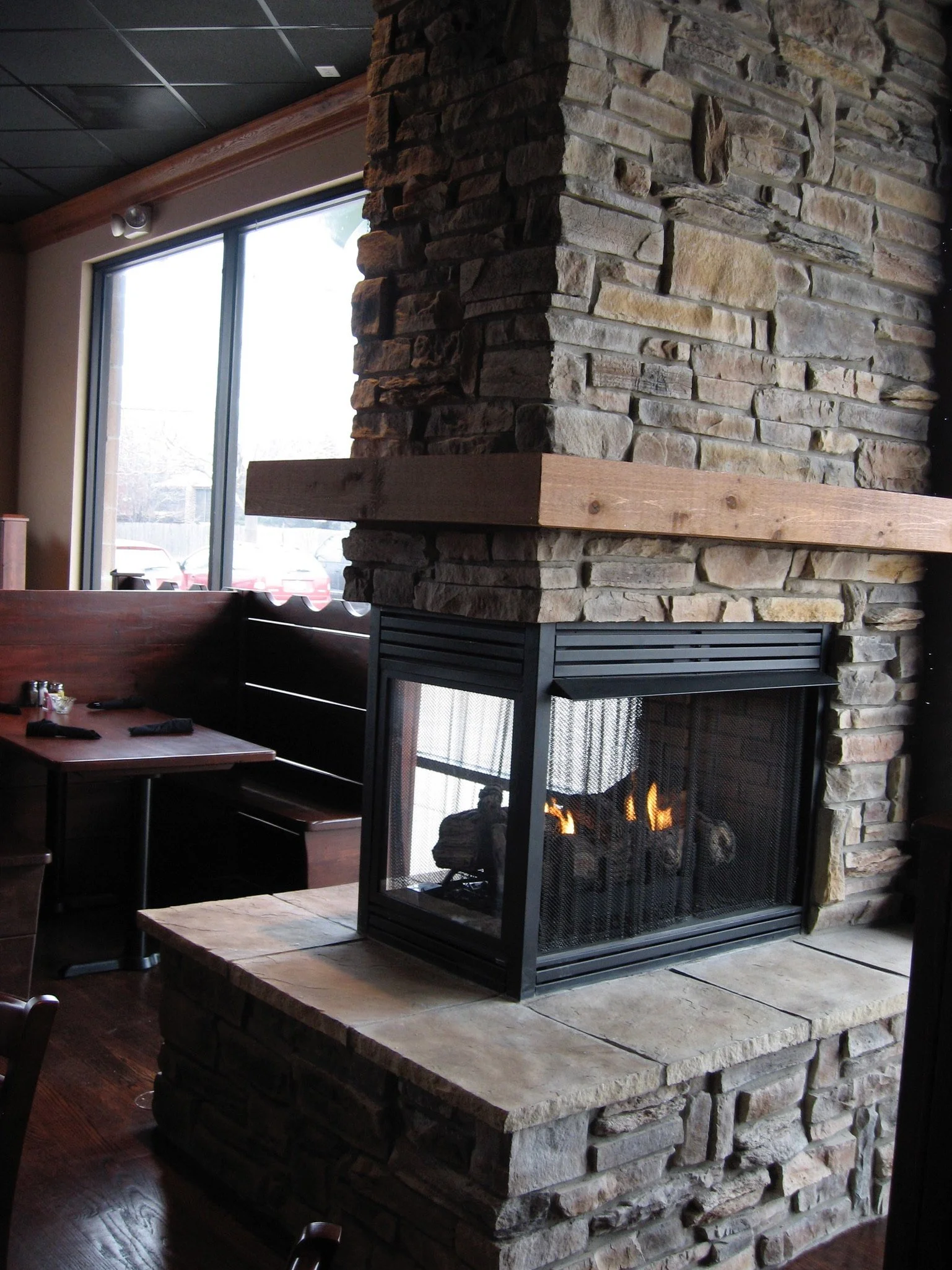 A cozy indoor space featuring a stone fireplace with a wood mantel, lit with a small fire, near a large window letting in natural light, with wooden furniture and flooring.