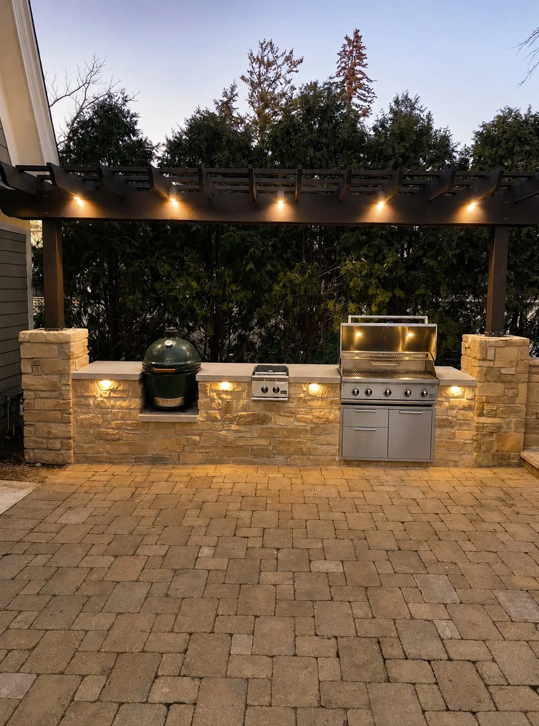Outdoor kitchen with stone counter, built-in grill, pizza oven, and a shaded pergola with string lights, set on a brick-paved patio, with trees in the background.