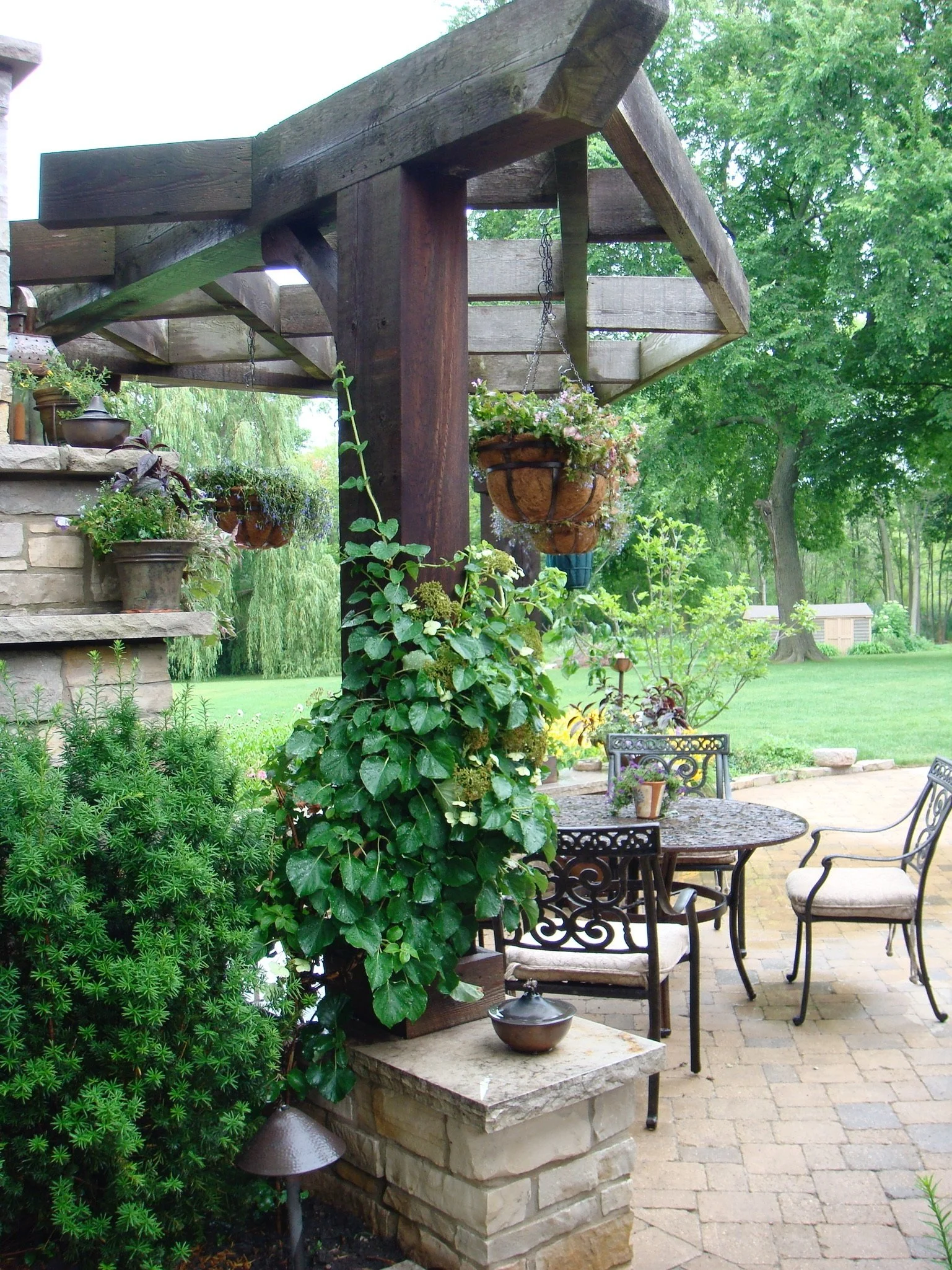 A backyard patio with metal outdoor furniture including a round table and chairs, surrounded by lush green plants, trees, and hanging flower baskets under a wooden pergola.