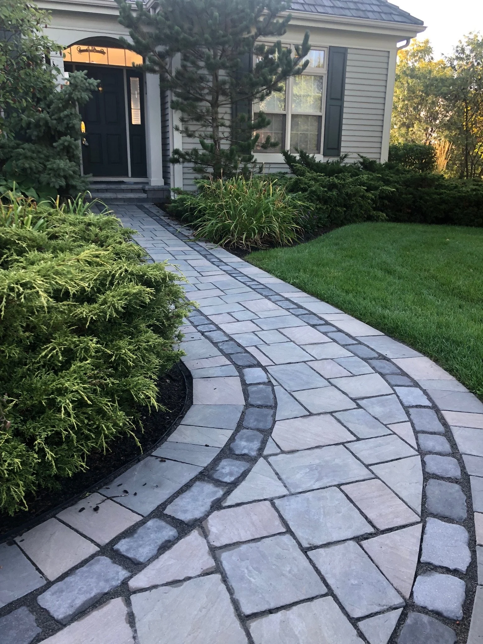 A stone pathway with dark grouting, winding through a landscaped yard with bushes, grass, and trees, leading to a house entrance with steps and a dark door.