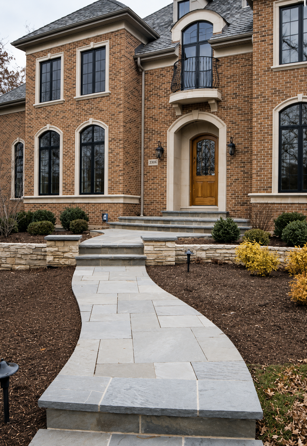 A brick house with tall black-framed windows, a curved front porch with stairs, and a small balcony above the entrance. The landscaped yard has a curving stone walkway and decorative shrubs.