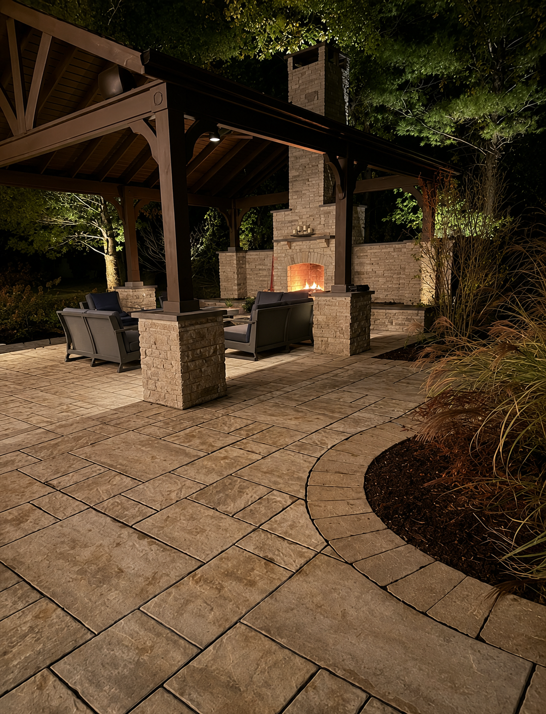 Nighttime outdoor patio area with stone flooring, seating, and a fireplace under a wooden pavilion, surrounded by trees and garden plants.