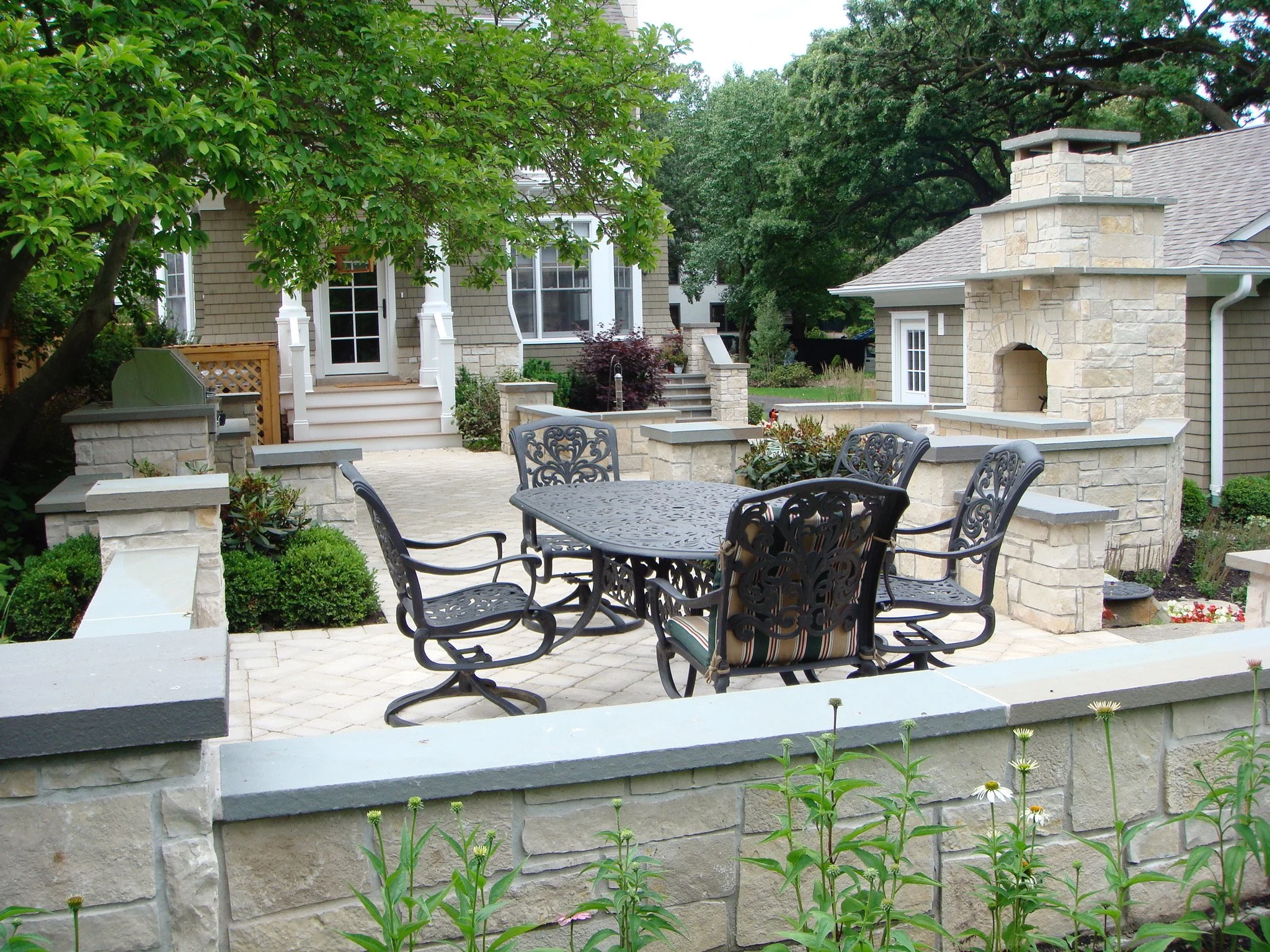A backyard patio area with a round metal table and six decorative chairs, surrounded by a low stone wall and lush plants, trees, and a house in the background.