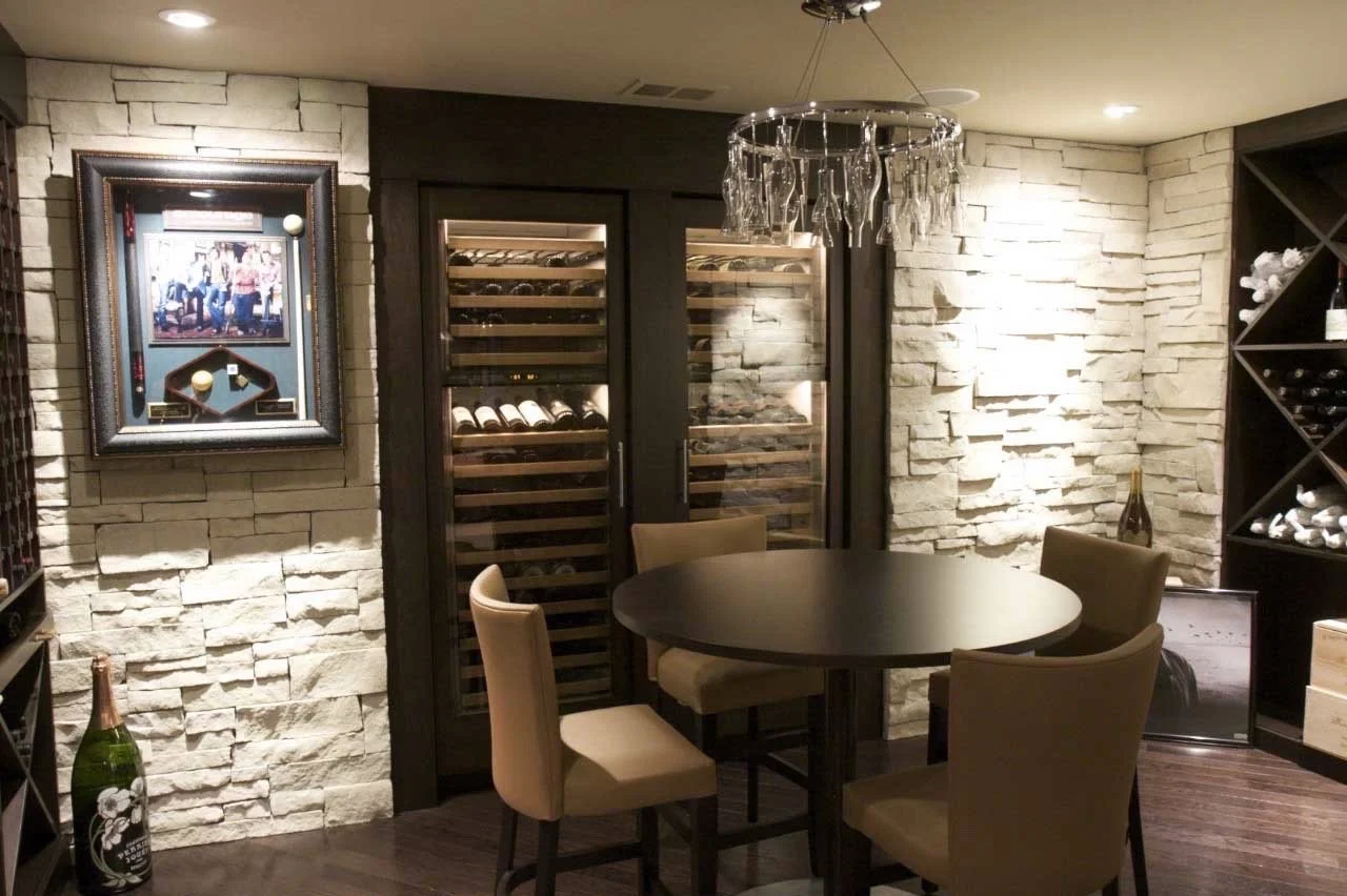 A wine cellar room with a round table, four beige chairs, wine bottles in a wine fridge, wall-mounted wine racks, and framed artwork on a stone wall.
