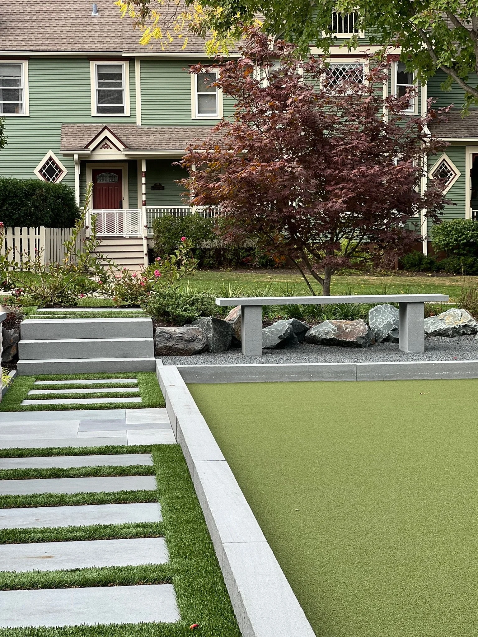 A well-maintained backyard with a small tree with reddish leaves, a white fence, garden bed with pink flowers, rocks, a staircase with grass on alternating steps, a wooden/metal bench, and an outdoor pool with a green cover. In the background, there 