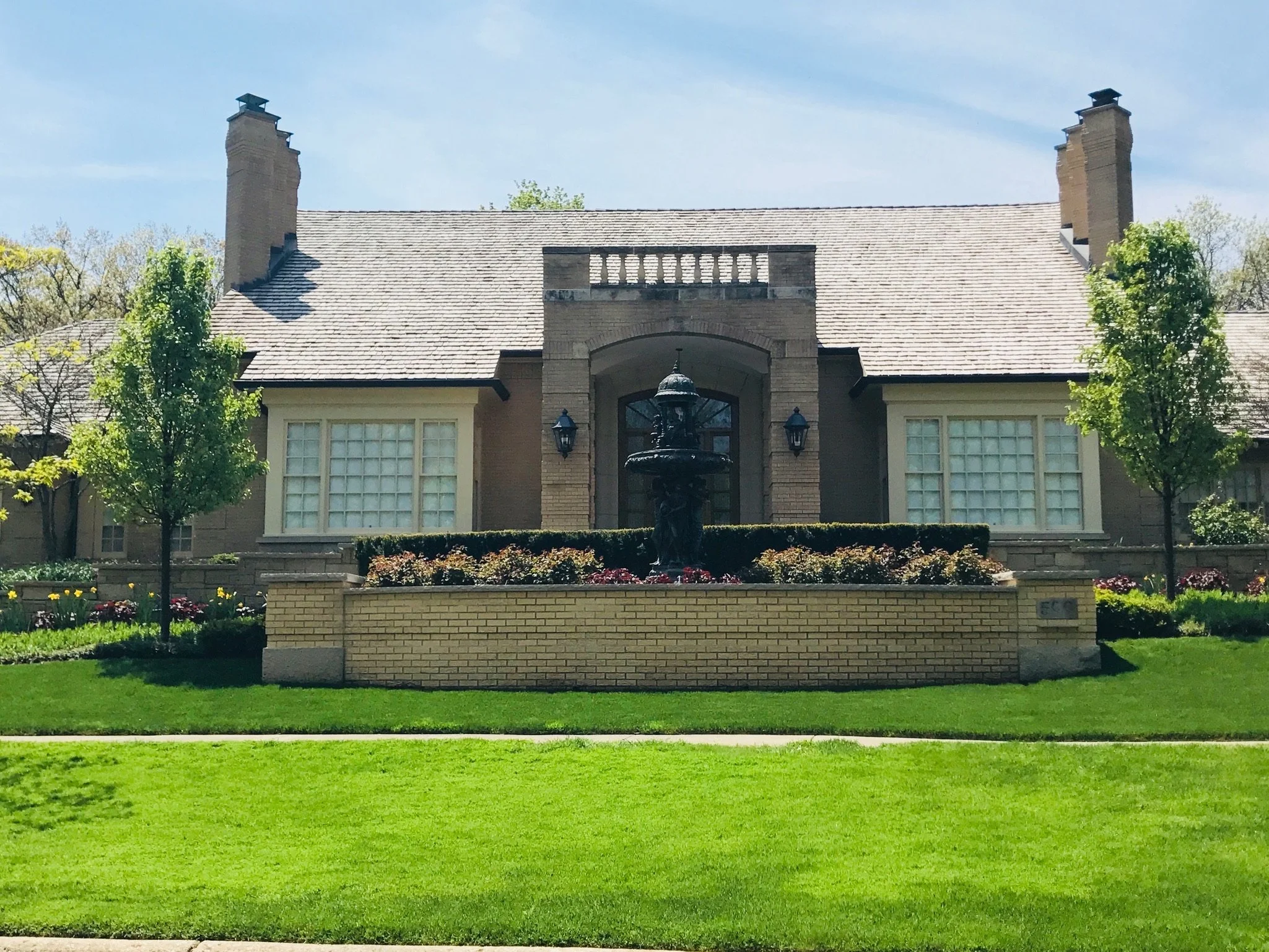 A large house with a steep, shingled roof, brick chimney stacks, and a front entrance featuring a fountain and decorative lighting. Surrounded by trees and a garden with flowers and neatly trimmed bushes.