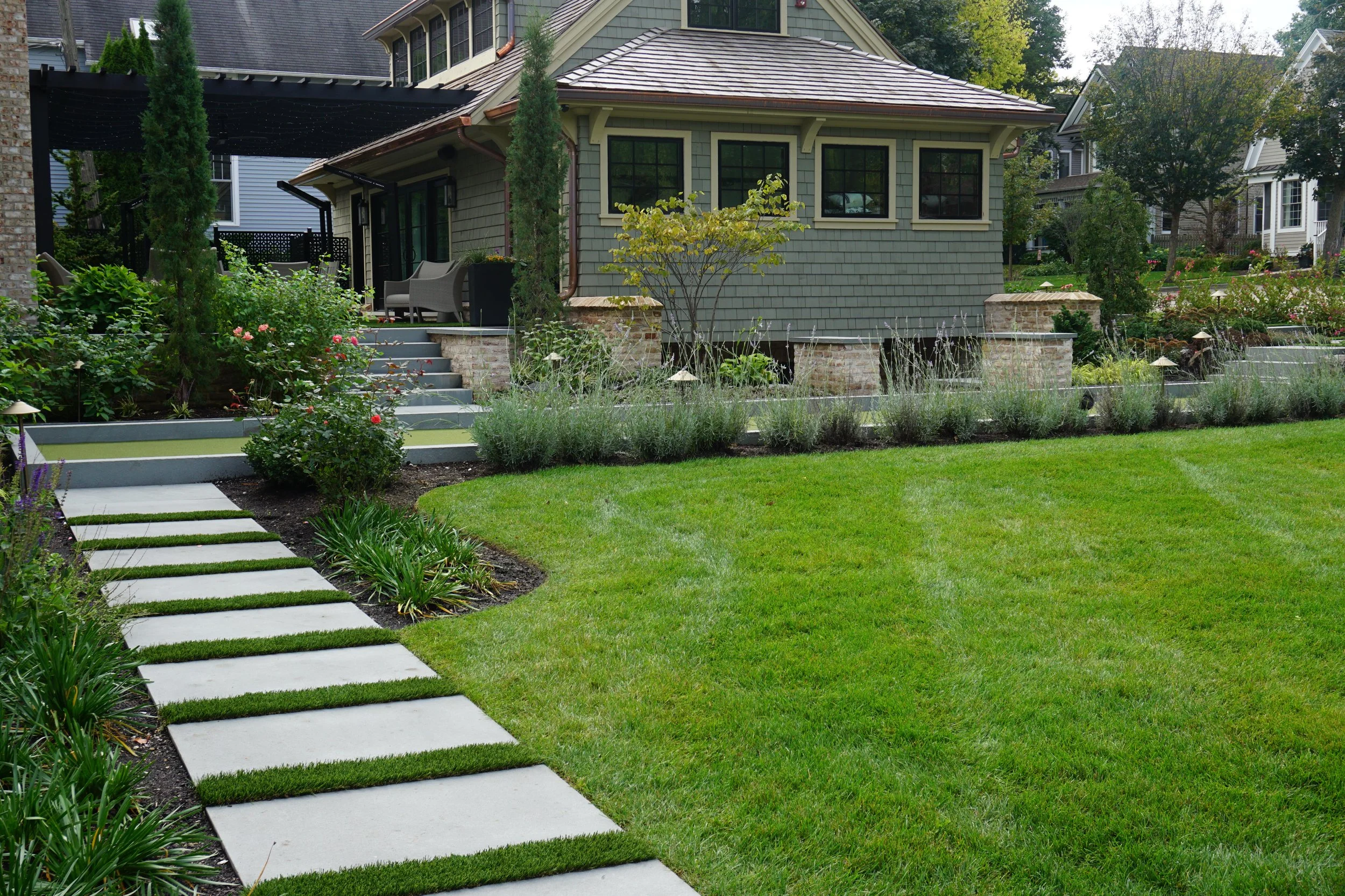 A well-maintained backyard garden with a grassy lawn, a pathway of stepping stones, flower beds, small trees, and outdoor seating near a house with green siding and black-framed windows.
