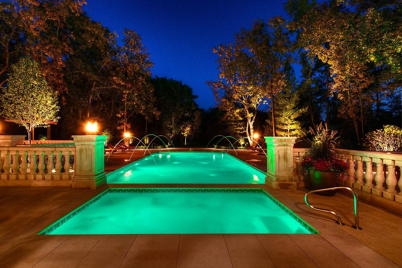 A beautifully lit swimming pool at night, surrounded by a stone terrace, decorative balustrades, trees, and outdoor lighting.