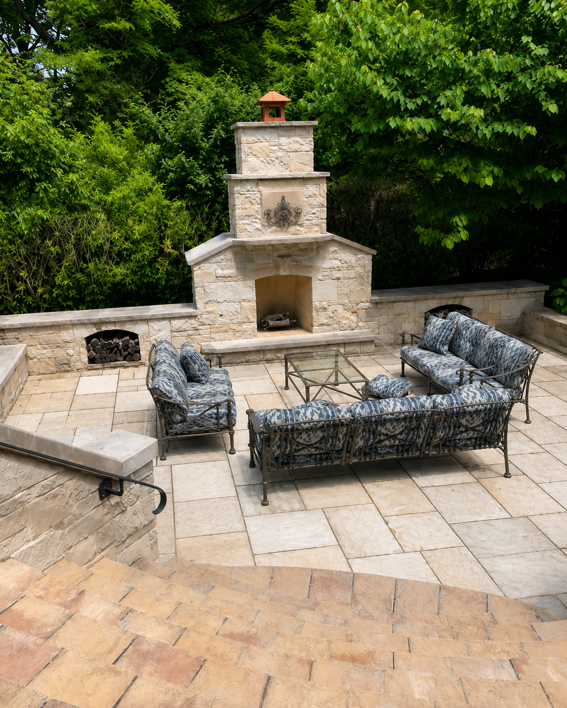 Outdoor patio with stone fireplace, glass-top coffee table, and outdoor seating with blue patterned cushions, surrounded by greenery.