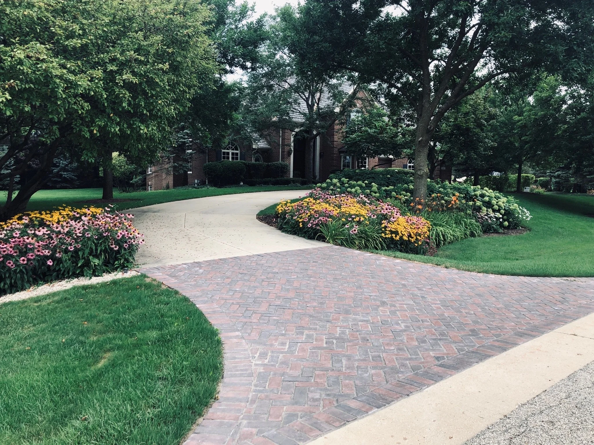 A brick pathway curves through a well-maintained lawn, lined with colorful flowers, leading to a brick house partially visible behind trees in a lush garden.