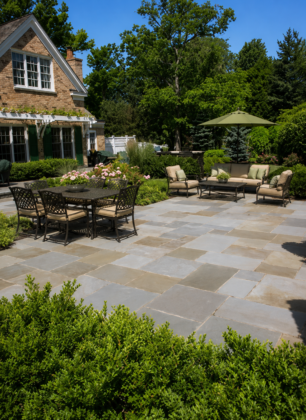 Spacious backyard patio with outdoor furniture, including a dining table with six chairs, a cushioned seating area with an umbrella, and lush green landscaping surrounding a brick house with green shutters and white fence in the background.
