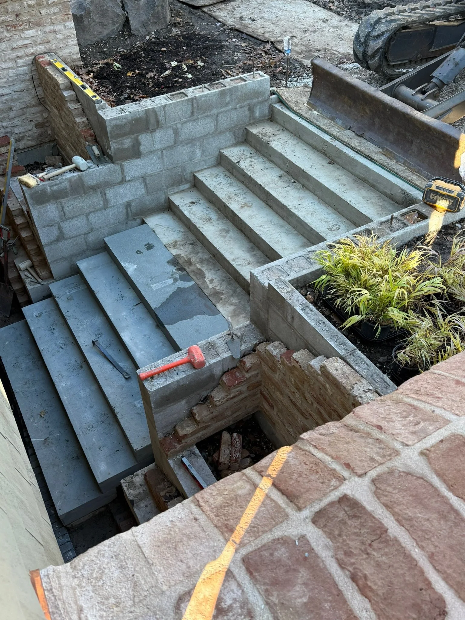 Construction site showing a set of concrete stairs being built, surrounded by construction tools, brick walls, and a small plants in a black container.