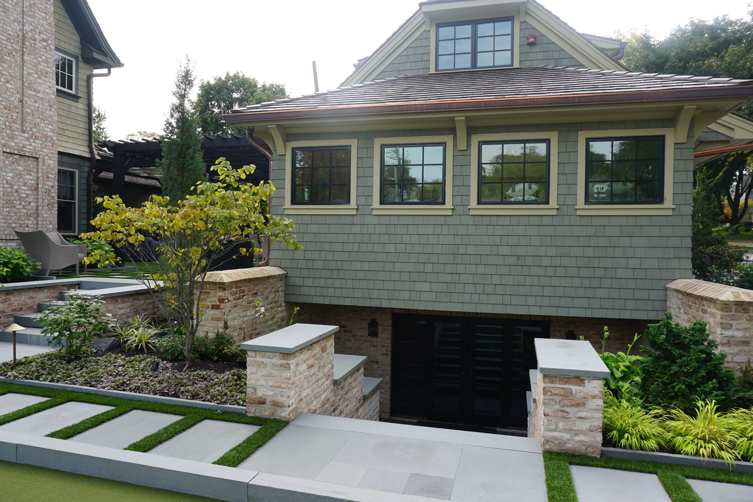 A backyard with a landscaped garden, patio area, and a house with green shingles and large windows. The patio has gray concrete slabs and a small grassy area with decorative plants. There is outdoor furniture and a tree with yellow-green leaves.