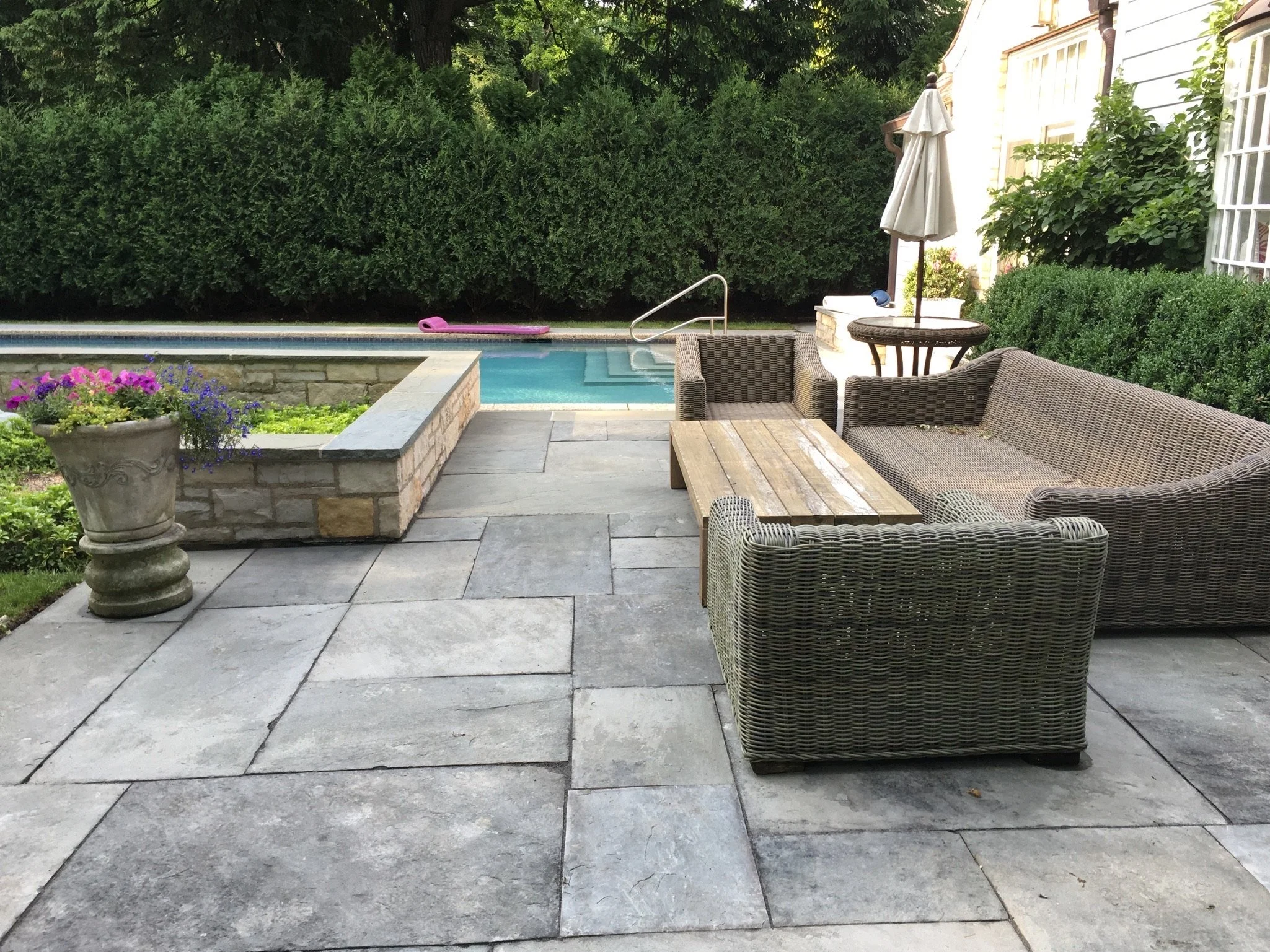 Backyard with a swimming pool, outdoor furniture including a wicker chair and table, an umbrella, colorful flowers in a pot, and dense green bushes and trees in the background.