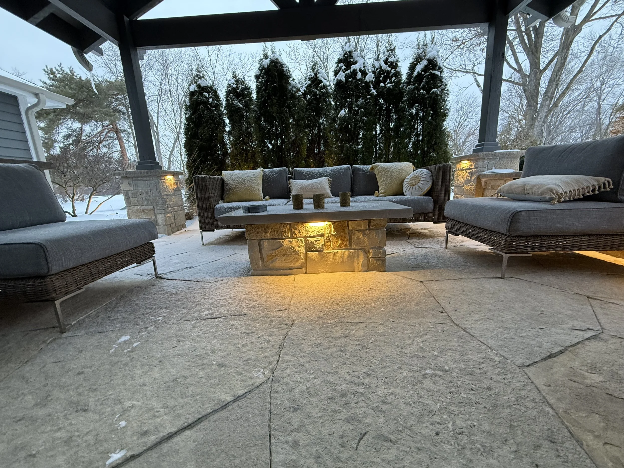 Covered outdoor patio area with cushioned seating, a fireplace, and snow on the furniture and ground, with trees in the background.