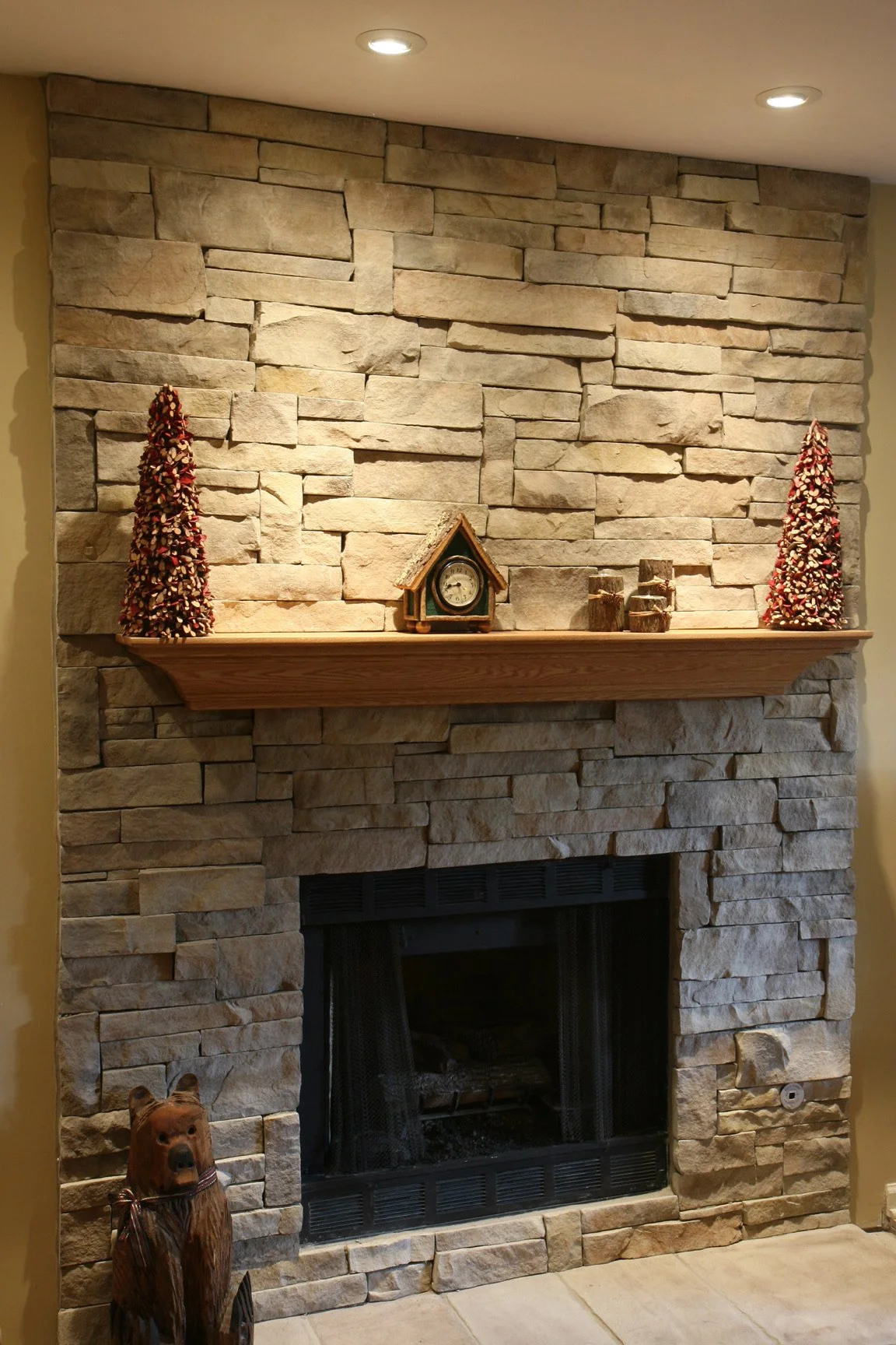 Decorative stone fireplace with a wooden mantel, adorned with two small artificial trees, a clock in a wooden house-shaped frame, and some wood log decorations. A carved wooden bear statue is positioned in front of the fireplace.