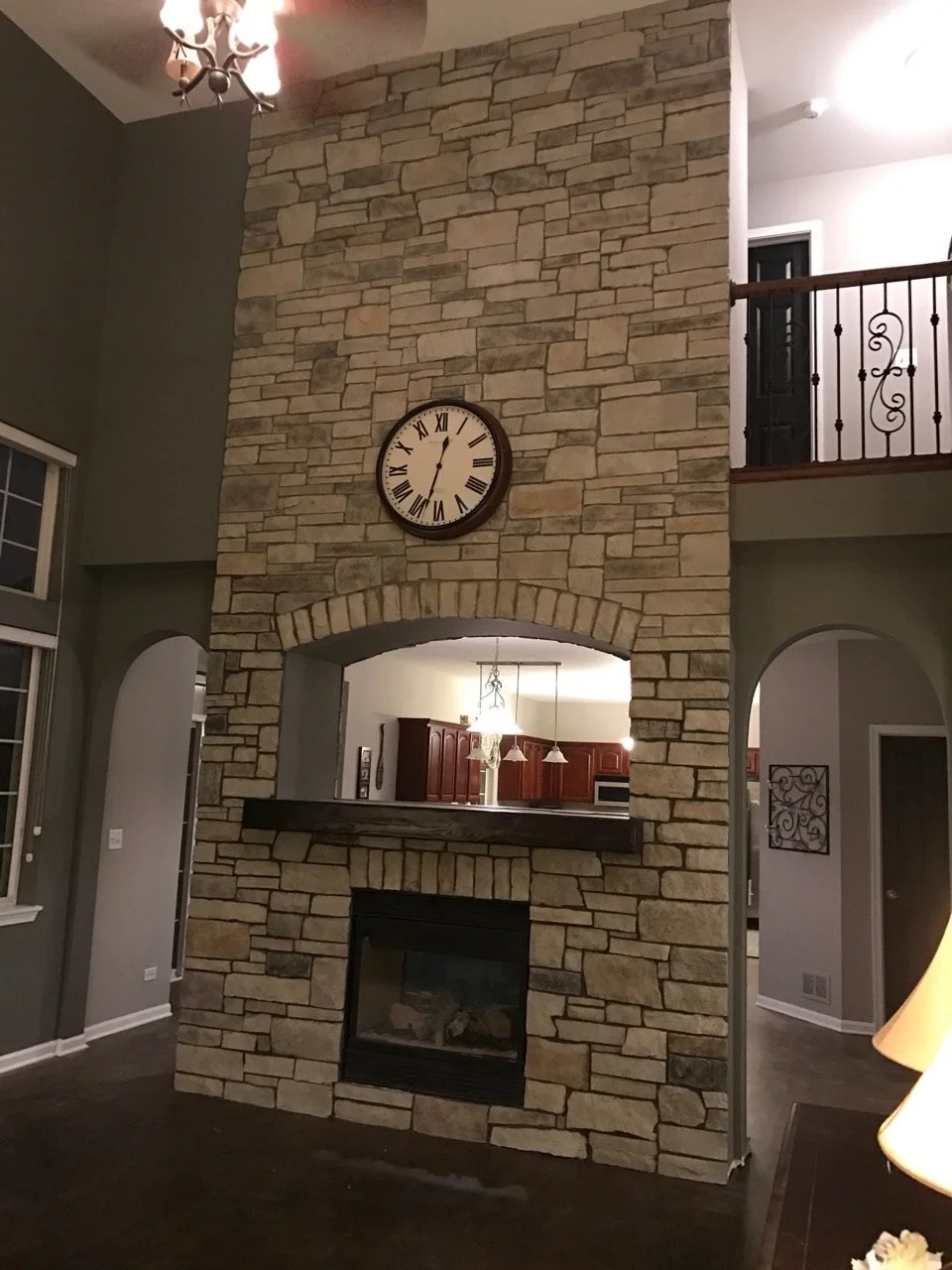 A tall stone fireplace with a clock mounted above it, and a cutout ledge across the middle of the fireplace. The kitchen is visible through the opening in the fireplace wall, with pendant lights hanging over a kitchen island.