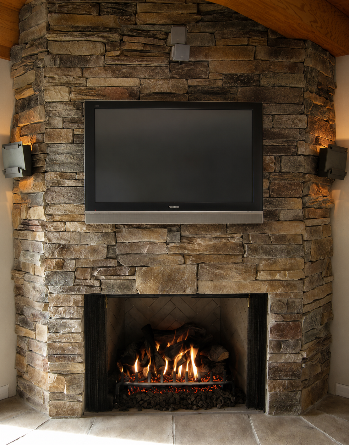 Indoor fireplace with a stone surround and a mounted flat-screen television above it, with two wall speakers on either side and a wood-paneled ceiling.