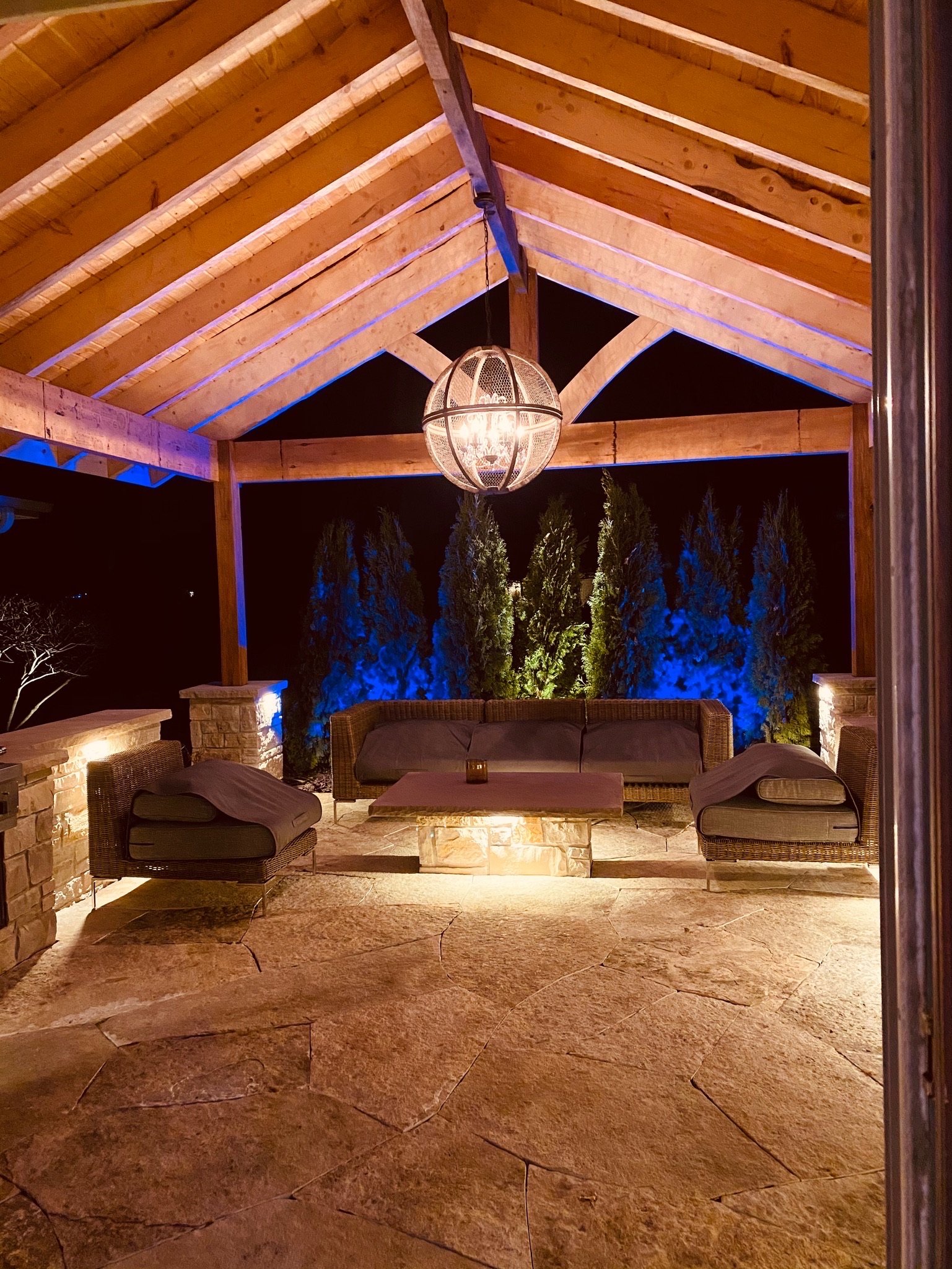 Nighttime outdoor patio with wooden roof, chandelier, wicker and cushioned seating, stone flooring, and blue accent lighting on trees.