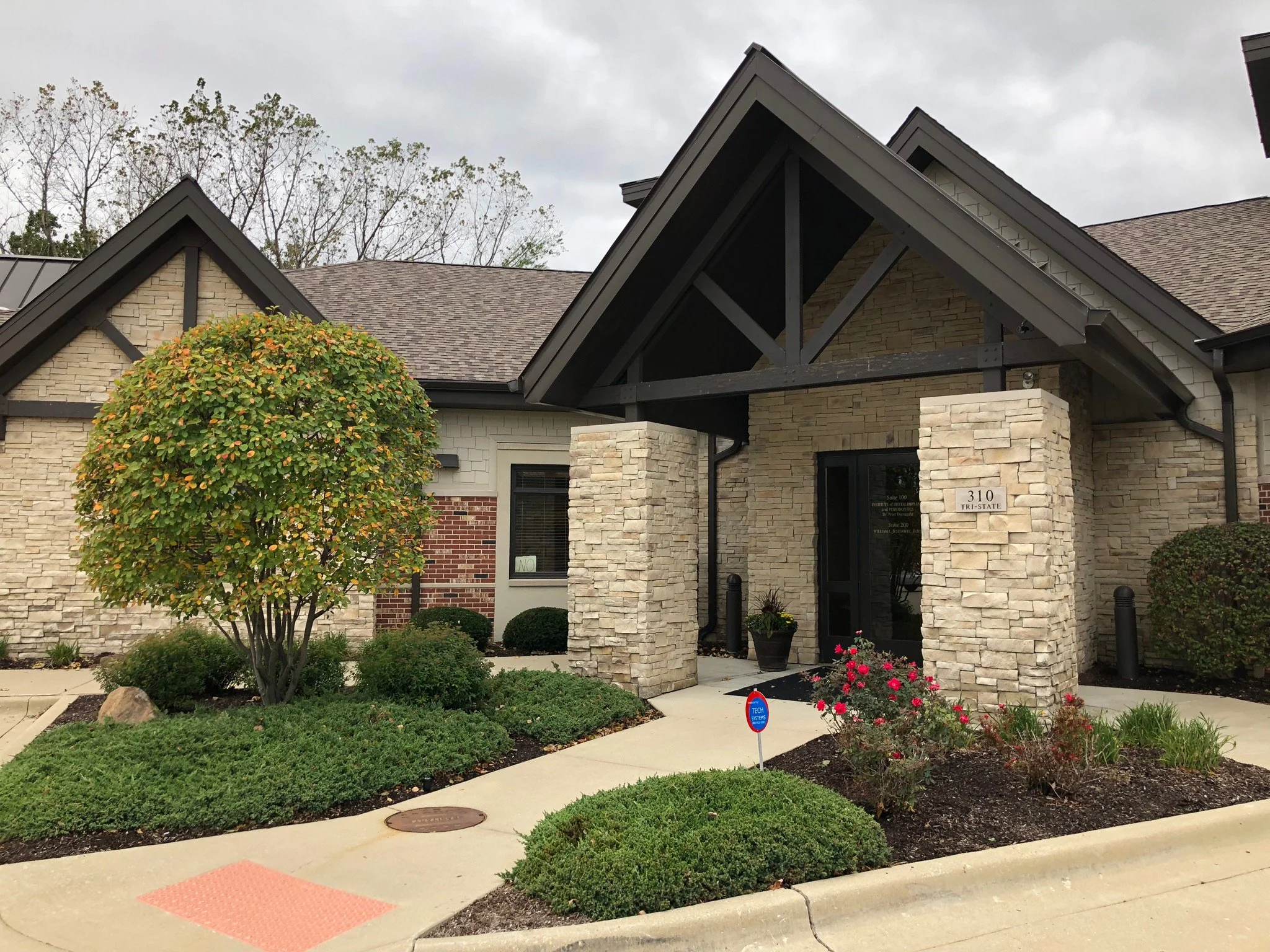 A modern house with beige brick walls, black trim, and a gabled roof. The front yard has a neatly maintained garden with a small tree, bushes, and flowering plants. A sidewalk runs in front of the house, with a red textured patch and a security sign.