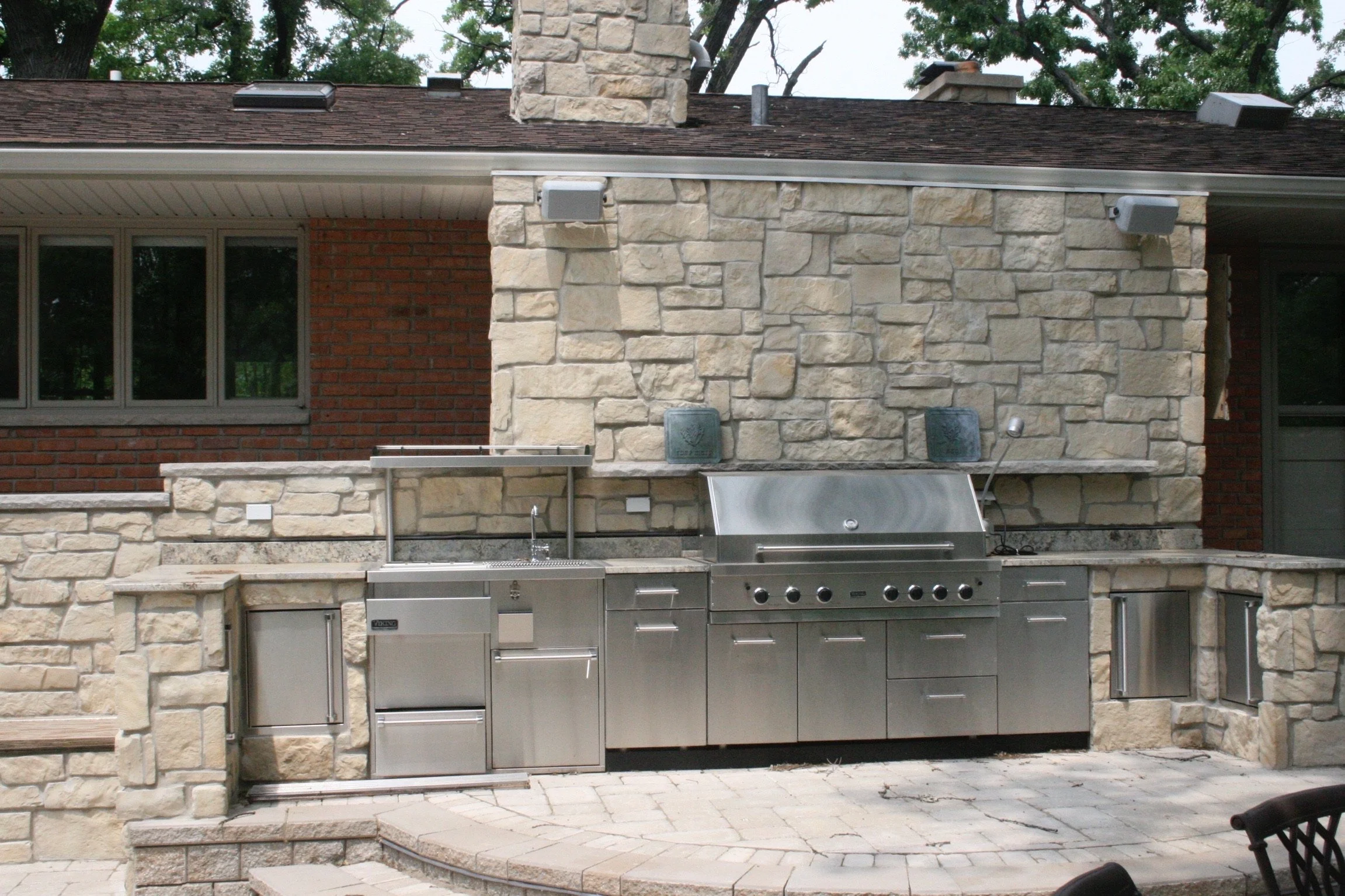 An outdoor kitchen with a stone and brick wall, metal grill, and stainless steel countertop under a roof, with trees in the background.