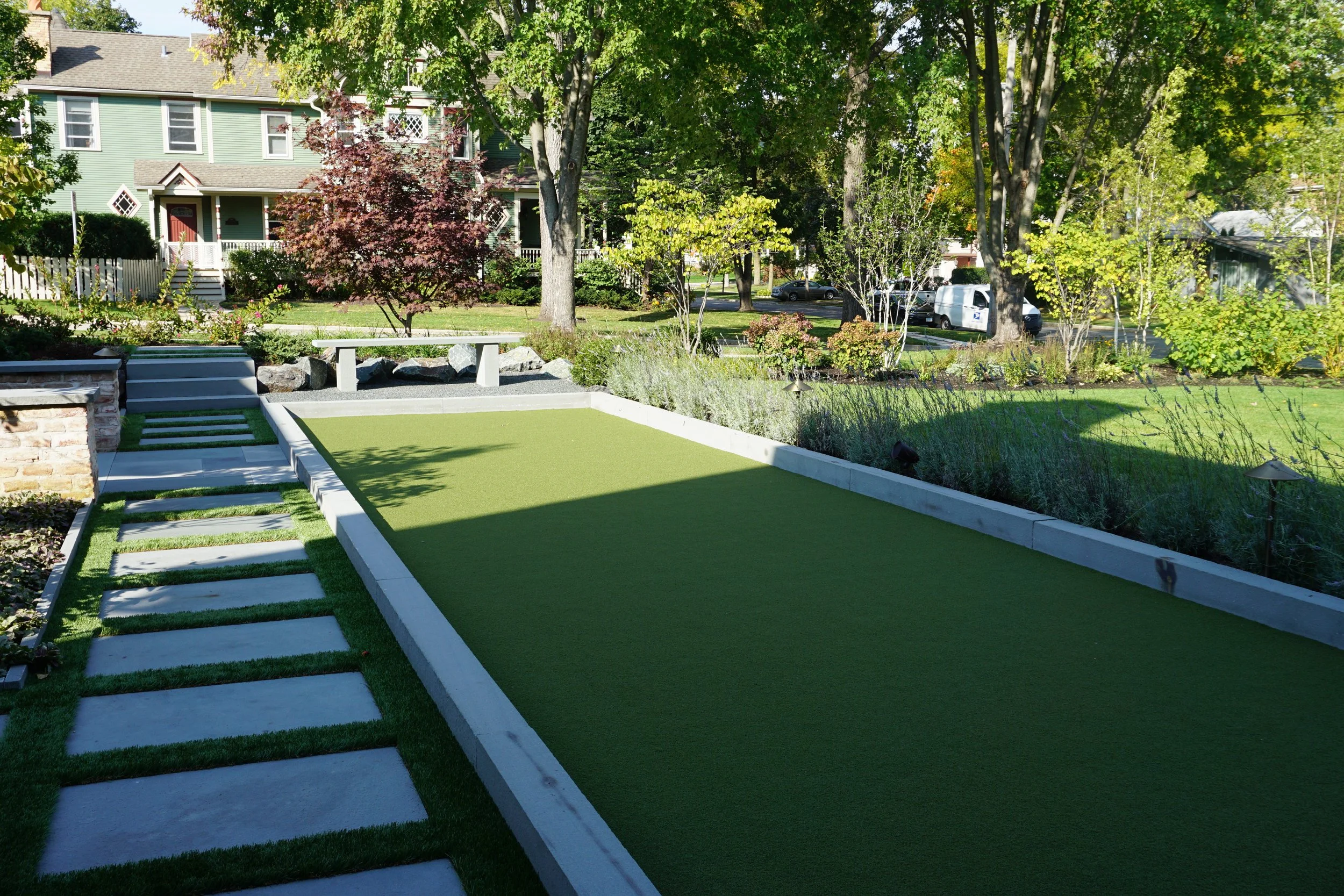 A backyard yard with a small artificial grass putting green, stone border, white pathway, and surrounding plants and trees. Residential houses and parked cars are visible in the background.