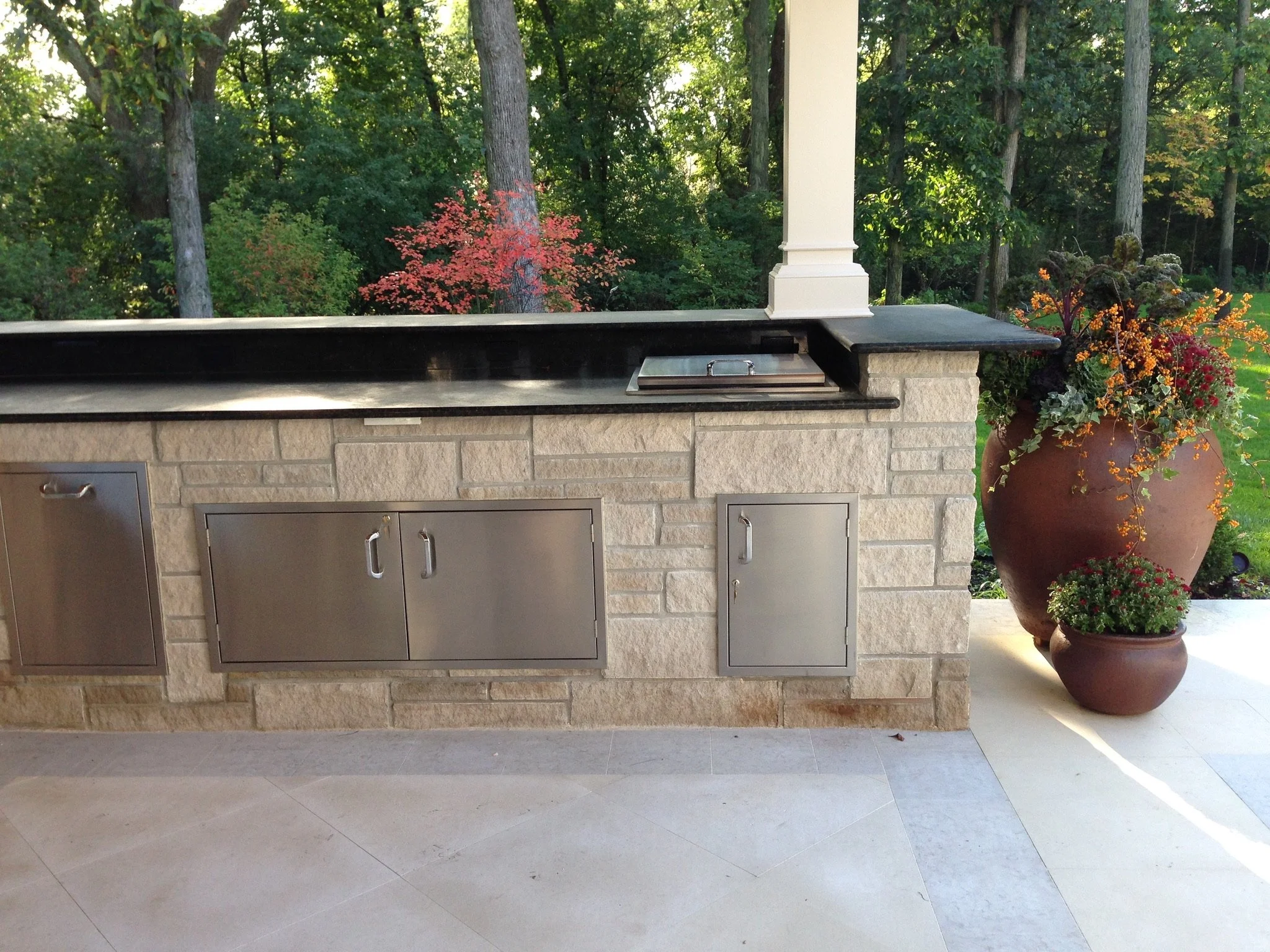 Outdoor kitchen with a stone counter and metal cabinets, decorated with large potted plants containing flowers, with a wooded background of trees and greenery.