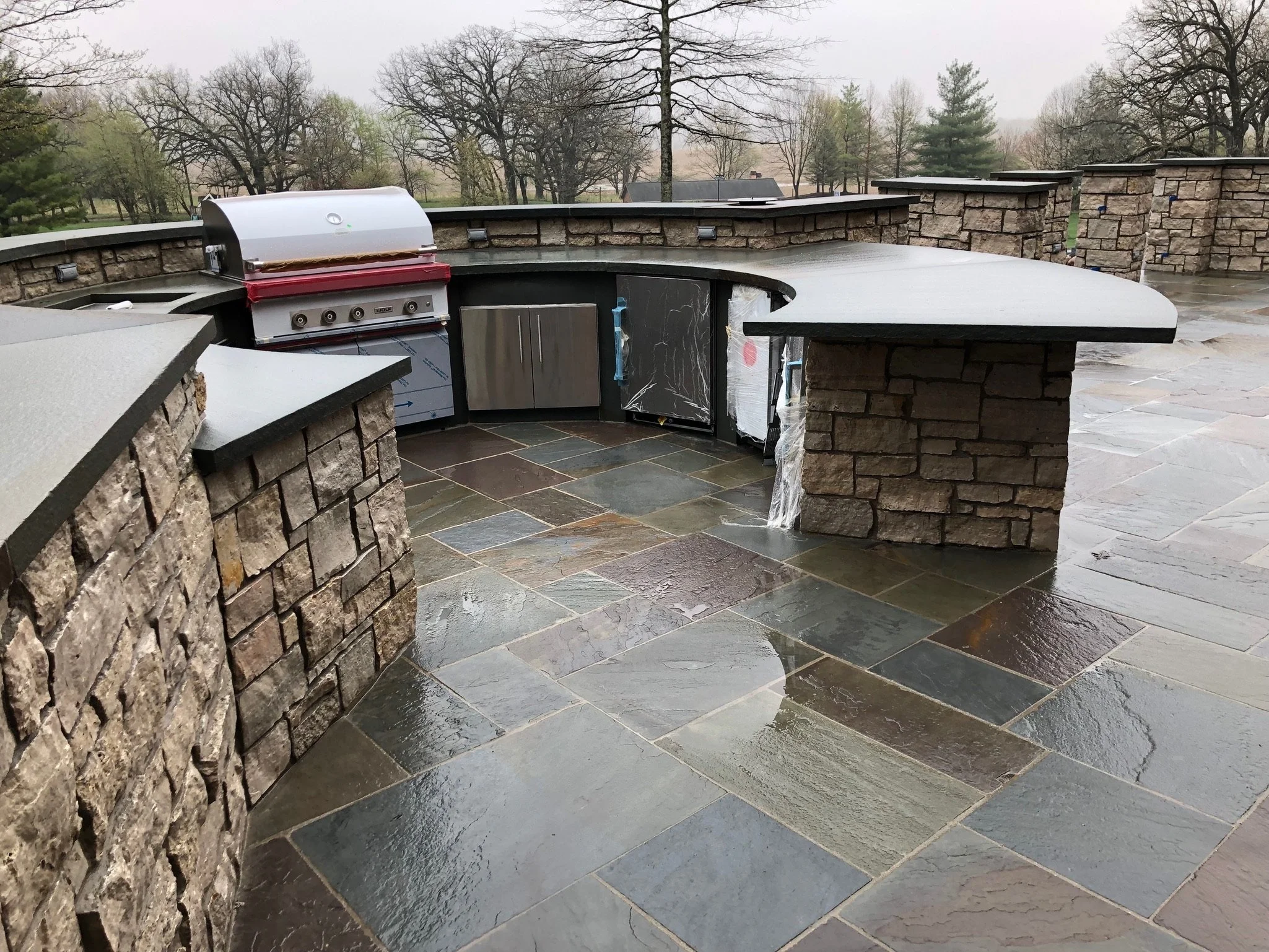Outdoor kitchen with stone counters, a built-in grill, stone island, and trees in the background on a cloudy day.