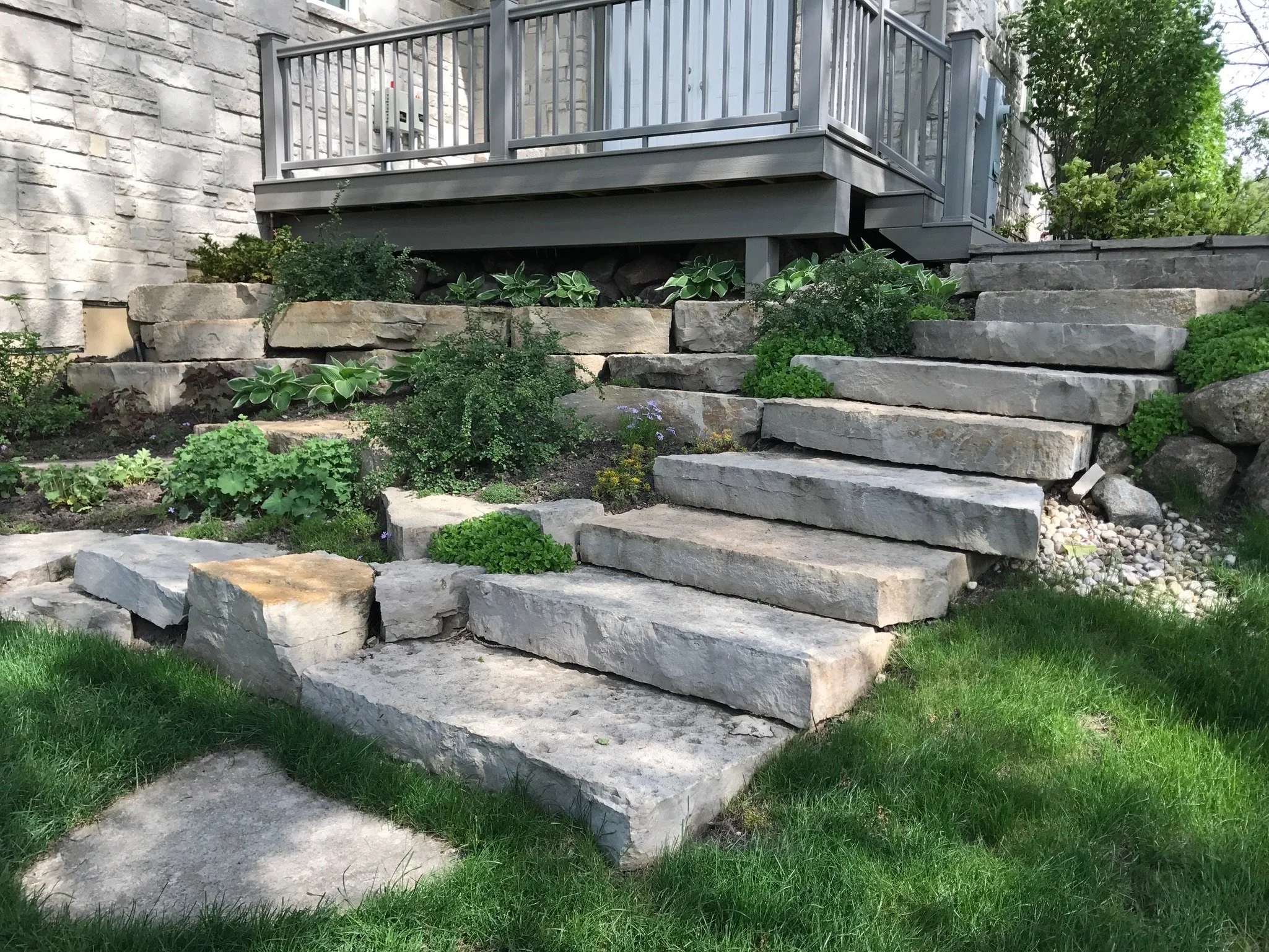 Stone steps leading up to a small porch with a gray railing, surrounded by green plants and a grass lawn.